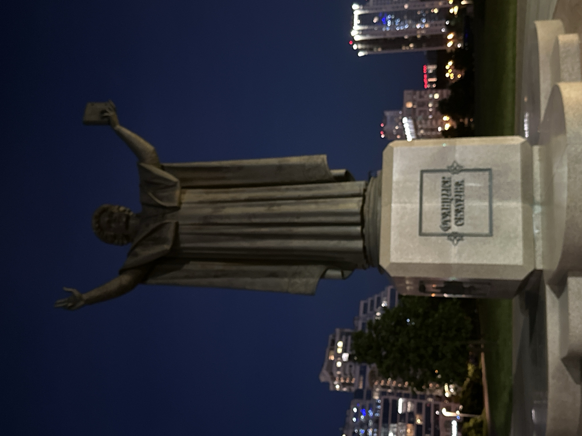 The image shows a statue of Francysk Skaryna in Minsk, Belarus, at night. The statue is bronze and depicts Skaryna holding a book aloft in one hand and extending the other hand outwards. The statue stands on a stone pedestal with an inscription in Belarusian Cyrillic script. In the background, blurred lights of high-rise buildings are visible against the dark blue evening sky. There is also greenery near the base of the statue, indicating a park or garden setting.