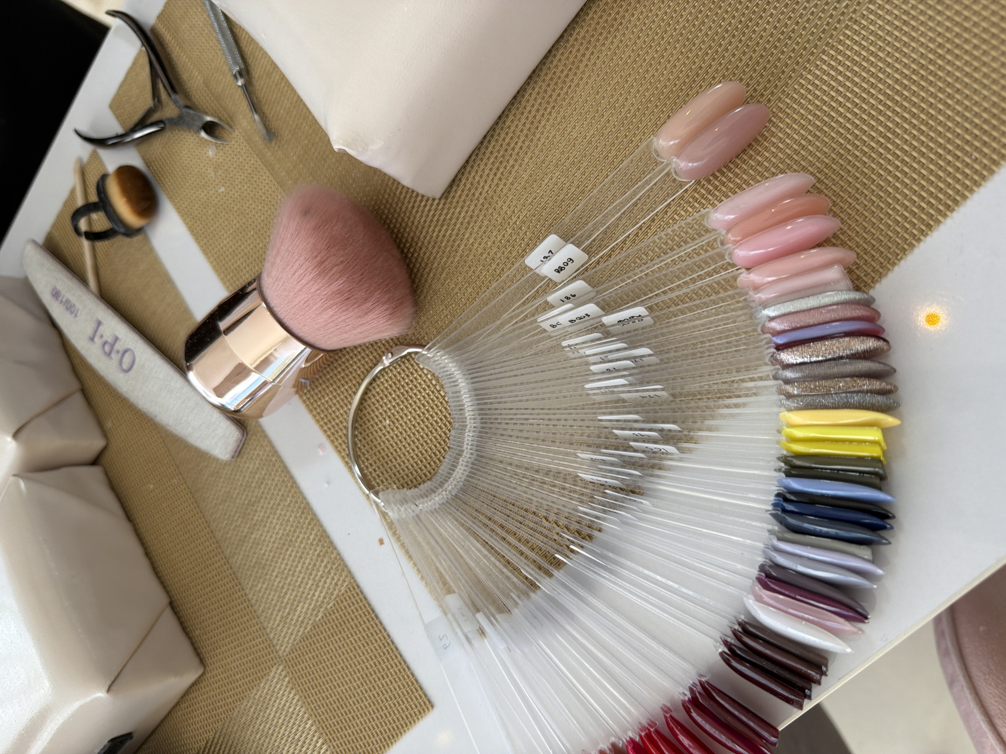 The image depicts a close-up of nail art supplies and tools arranged on a tan-colored, textured surface with white stripes, possibly a manicure station. The scene is located in North Kuta, Indonesia.
Objects present include:
* A nail polish color wheel with a range of colors from pinks and nudes to reds, purples, blues, yellows, and glittery shades. Each nail tip on the wheel has a corresponding label attached to its base.
* A large, pink-bristled makeup brush with a gold handle.
* A white OPI-branded nail file.
* A small black dappen dish with a brown substance inside and a black ring surrounding it.
* A set of cuticle clippers and a cuticle pusher.
* A small white cushion, possibly for hand support.
The scene appears to be a workspace for nail services, likely in a salon or studio. The arrangement suggests a focus on selecting nail polish colors.