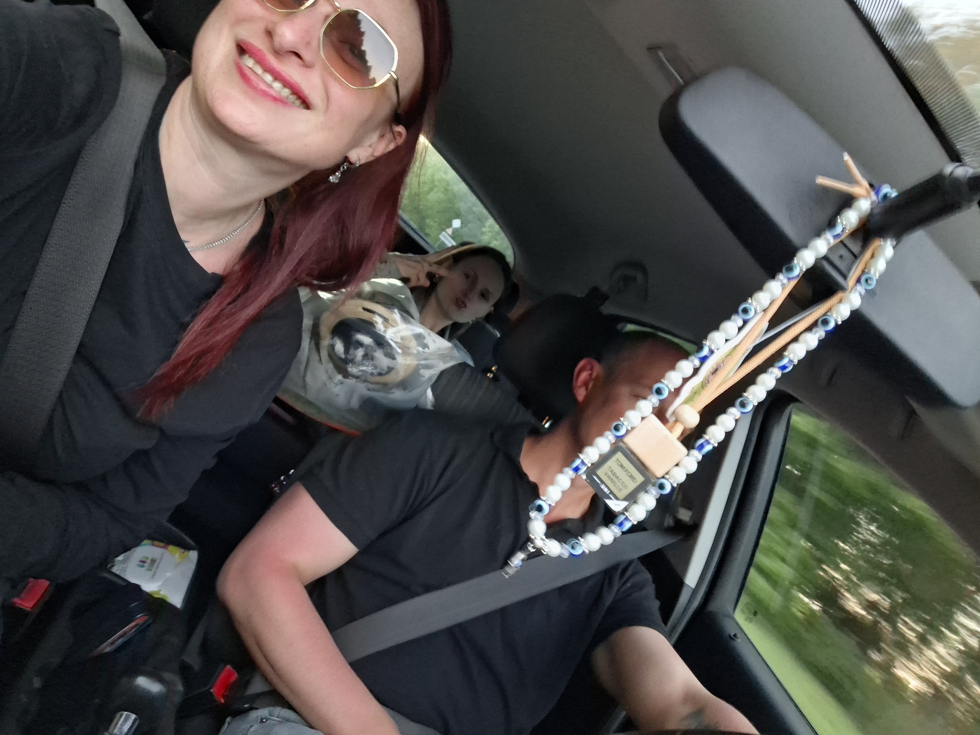 Captured from inside a moving vehicle, the image shows three individuals and various objects in Любанскі сельскі Савет, Belarus.
The foremost person, on the left, is a woman wearing sunglasses, a black shirt, and a seatbelt. Her hair is dyed a deep red. In the back seat, another woman is visible. She has a clear plastic bag next to her. In the driver's seat is a man also wearing a black shirt and a seatbelt.
Hanging from the rearview mirror is a beaded car accessory, adorned with eye-shaped charms and a perfume tag that reads "TOM FORD TABACCO VANILLE." The vehicle's interior is visible, including the dashboard and door panel. Outside the car, blurred trees suggest the car is in motion.