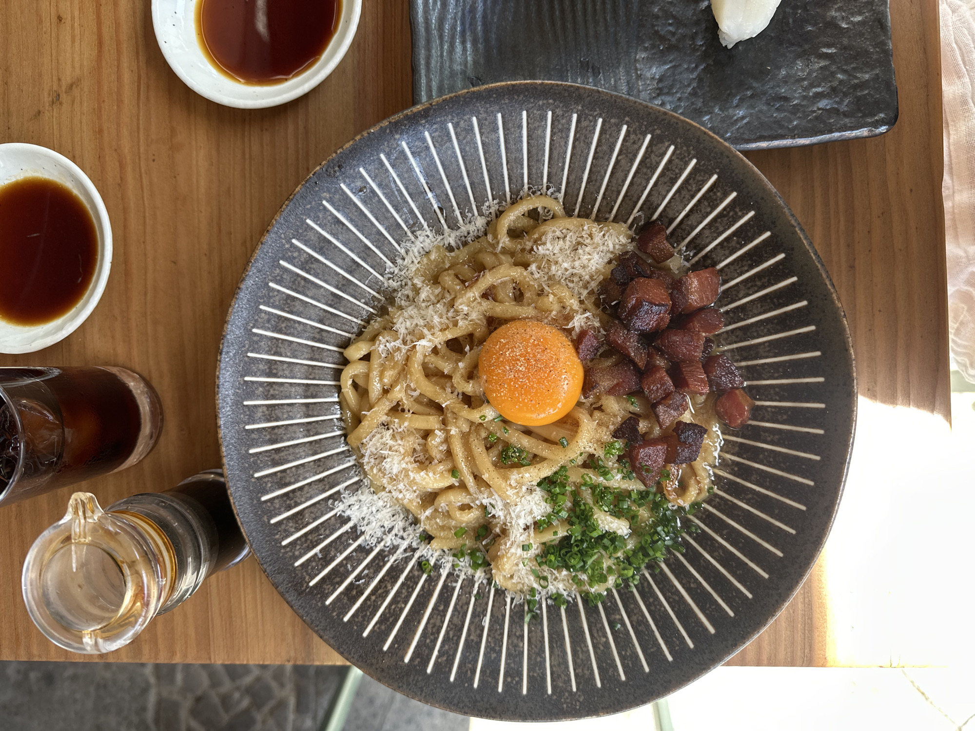 An overhead shot shows a plate of Mazemen-style noodles on a wooden table. The noodles are garnished with shredded cheese, an egg yolk, chives, and diced meat (possibly pork). Two small bowls of soy sauce and a glass of dark liquid (likely iced coffee) are arranged nearby. The location is uncertain but might be in Berlin, Germany.