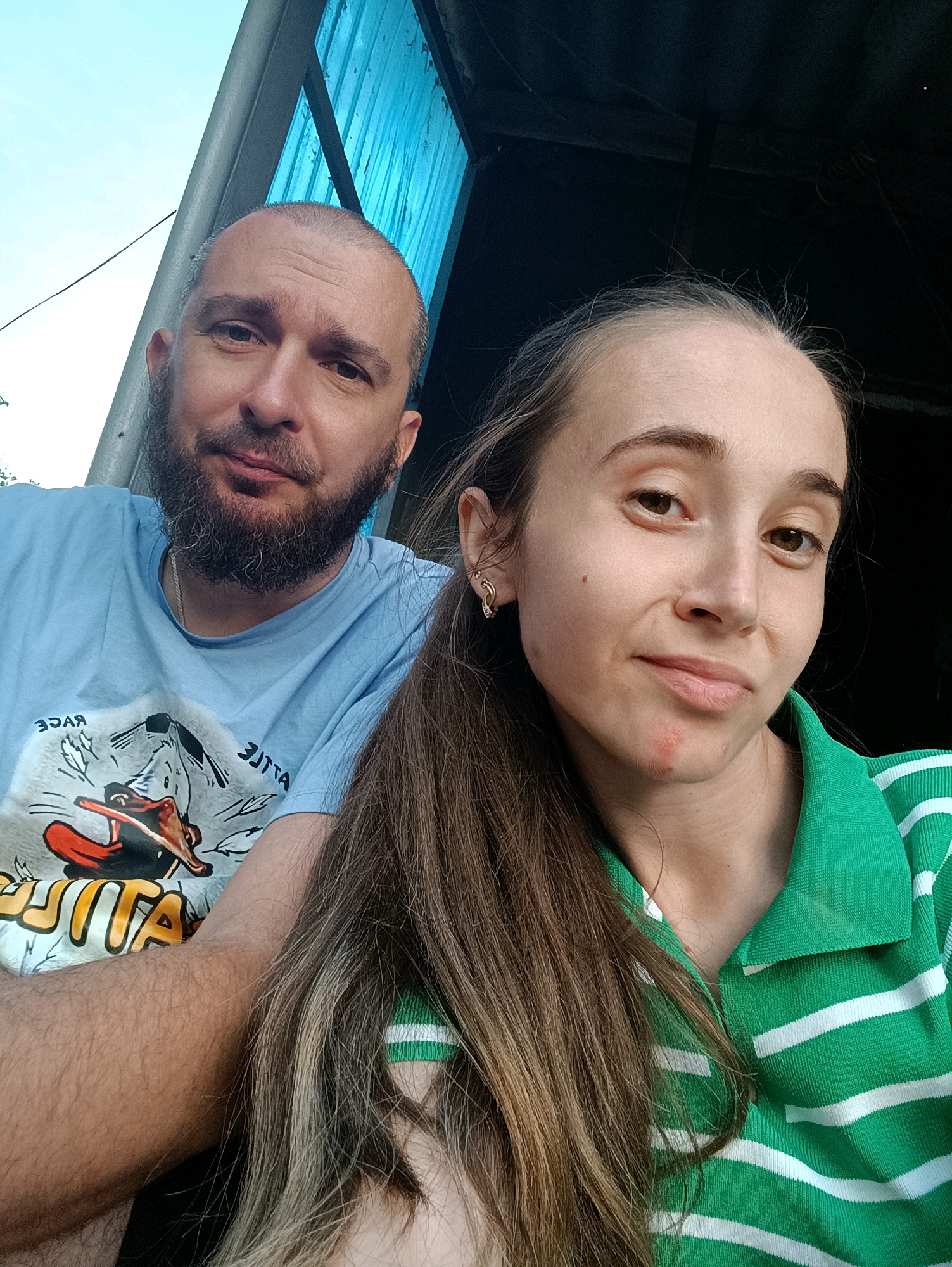 This is a selfie taken by two people. A man with a beard and a blue t-shirt is on the left, and a woman with long brown hair and a green and white striped shirt is on the right. They are in an outdoor location, possibly Vypasne, Ukraine, indicated by a blue corrugated plastic roof and a light pole in the background. The man is looking at the camera with a neutral expression, while the woman is smiling slightly. There is some text on the man's t-shirt that is partially obscured. The overall lighting suggests it is daytime.