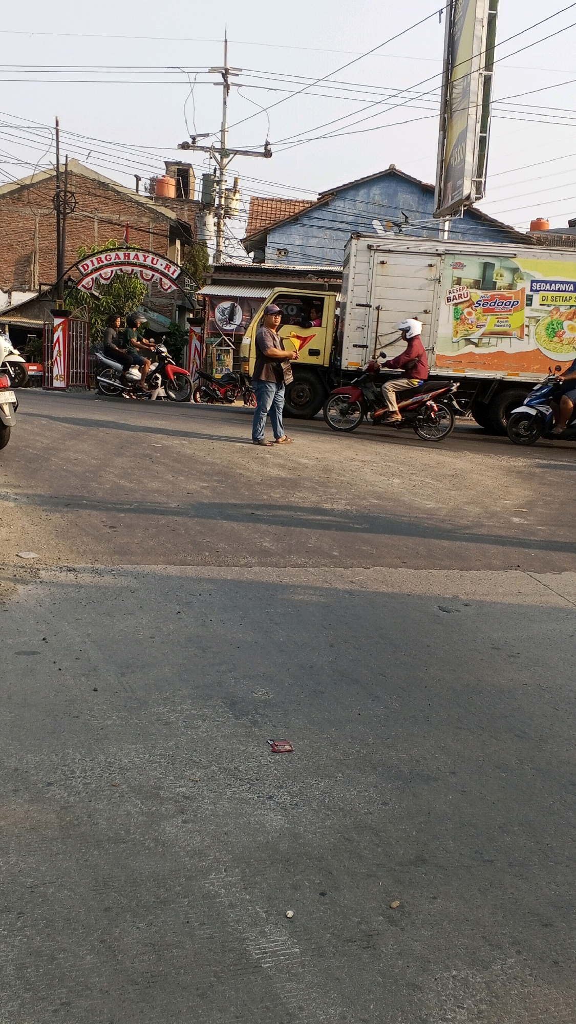 The image depicts a street scene in Semarang, Indonesia, featuring a busy intersection with several motorcycles and a delivery truck. The foreground shows a paved road with some debris. In the middle ground, a yellow delivery truck is parked on the right side of the road, displaying advertisements for instant noodles. A person on a red motorcycle wearing a white helmet is riding in front of the truck. To the left of the truck, a man wearing a cap and blue jeans is standing and holding a yellow object. Several motorcycles are parked on the left side of the street, with some individuals on them. In the background, buildings with red brick and blue siding are visible, along with utility poles and overhead wires. A decorative archway with the words "DIRGAHAYU RI" is also present. The overall scene suggests a typical urban environment in Semarang during daylight.