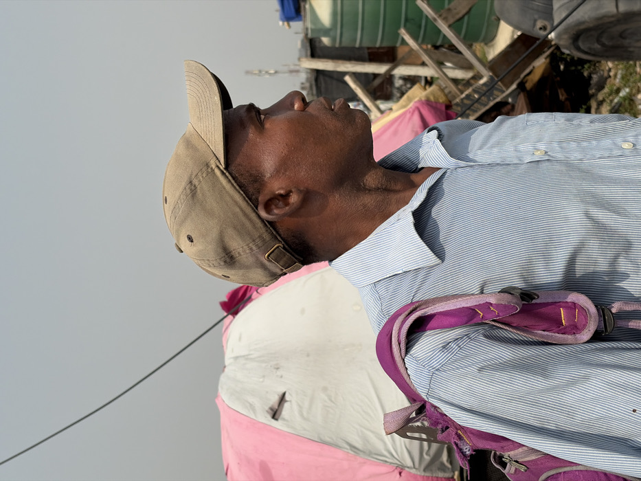 The image captures an adult male in Igboefon, Nigeria, depicted from the chest up in profile, facing towards the right. He has dark skin and wears a faded olive-brown baseball cap with a metal buckle at the back, along with a light blue pinstriped collared shirt. A purple backpack, showing signs of wear and fraying on the strap, is slung over his left shoulder. His mouth is slightly open. In the background to the left, a portion of a pink and white temporary shelter or tent is visible, exhibiting wear and marks. A thin, dark cable diagonally crosses the upper left quadrant of the frame. To the man's right, the background presents a cluttered outdoor environment, including parts of green cylindrical tanks, wooden debris, and additional textile materials. The sky is uniformly hazy light grey.