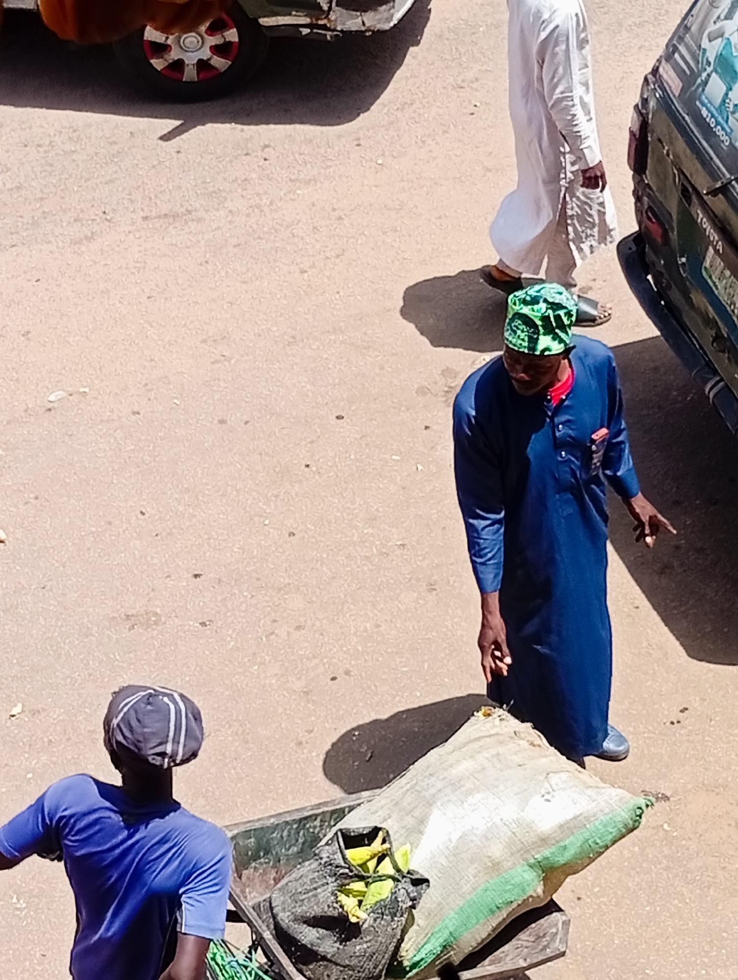 In Bukuru, Nigeria, two men are in a street scene. A man wearing a blue tunic and a green patterned cap stands near a dark vehicle. He is gesturing with his right hand. In the foreground, another man in a blue t-shirt and a dark cap with white stripes is pushing a cart that contains a large sack. The ground is dusty and uneven. A white robed figure is visible in the background, walking away from the camera.