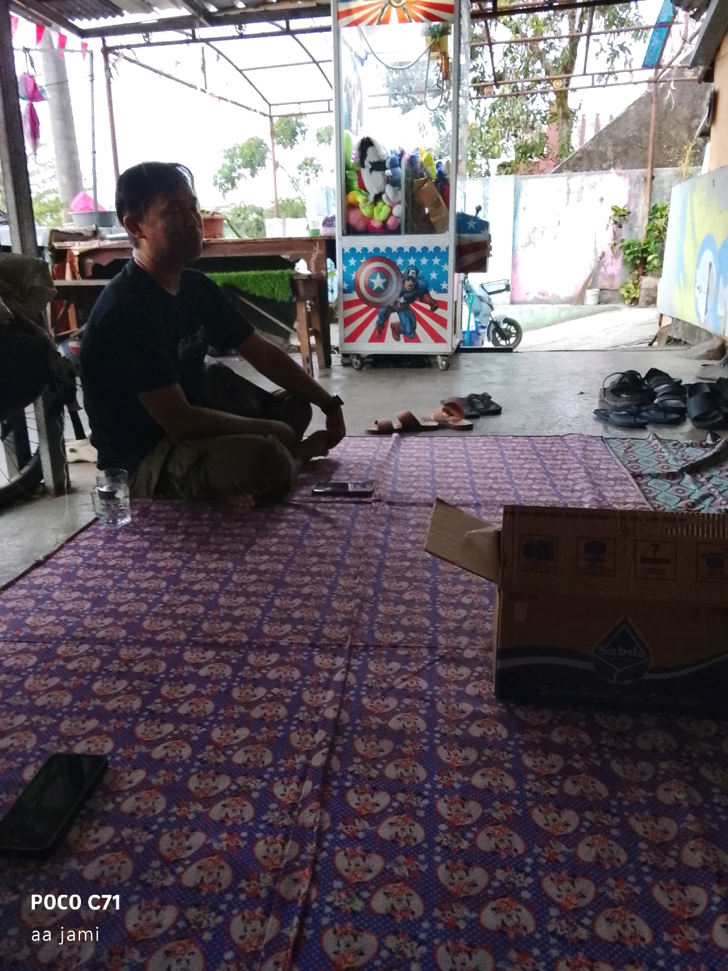 The image depicts an outdoor scene, possibly a backyard or a small open-air market stall, in Tasikmalaya, Indonesia. A man is seated cross-legged on a purple mat adorned with a cartoon pattern, facing away from the camera and looking towards the right. He is wearing a black t-shirt and light-colored pants. To his left, on the mat, is a glass of water. In the background, there is a red, white, and blue arcade machine, likely a claw machine, filled with stuffed toys. To the right of the arcade machine, a small, white scooter is parked. Further in the background, there are buildings with concrete walls and a tiled roof. Several pairs of sandals are scattered on the ground. A cardboard box is placed on a mat in the foreground, to the right. The overall impression is of a casual, possibly commercial, outdoor setting in an Indonesian city.