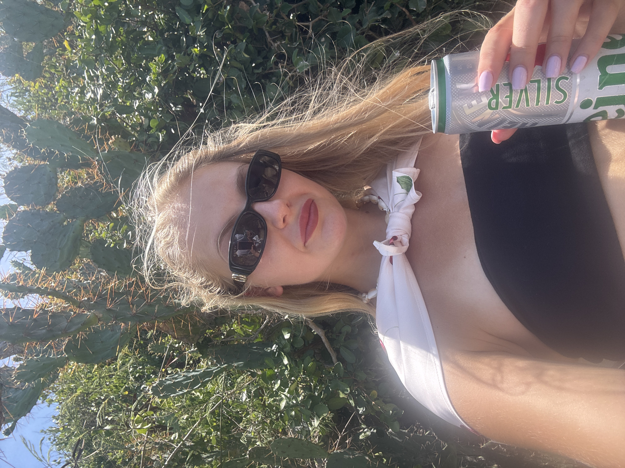 A woman with blonde hair and sunglasses is holding a silver can in her hand. She is wearing a black top and a white scarf with a floral pattern tied around her neck. Her fingernails are painted a light purple. The background is a natural setting with green bushes and cacti. The lighting suggests it is daytime, likely outdoors. The woman appears to be taking a selfie. The can has the word "SILVER" visible on it in green text.