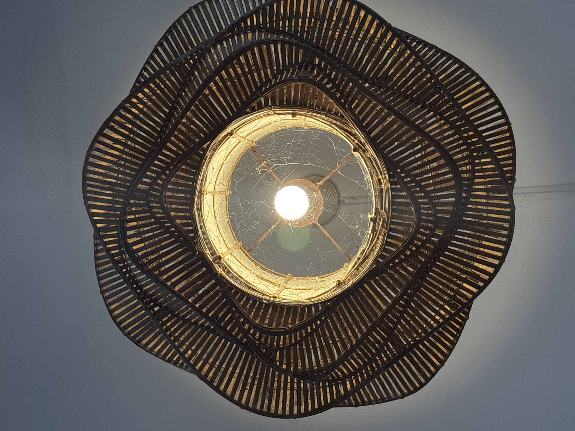This is an extreme close-up, overhead shot of a woven lampshade in Chalong, Thailand. The lampshade is made of dark, thin reeds or bamboo strips arranged in concentric, wavy layers that radiate outwards from a central light bulb. The light bulb emits a bright, warm glow, illuminating the woven texture of the shade and revealing a delicate spiderweb stretched across the inside of the shade, directly around the bulb. The background is a plain, light gray ceiling.