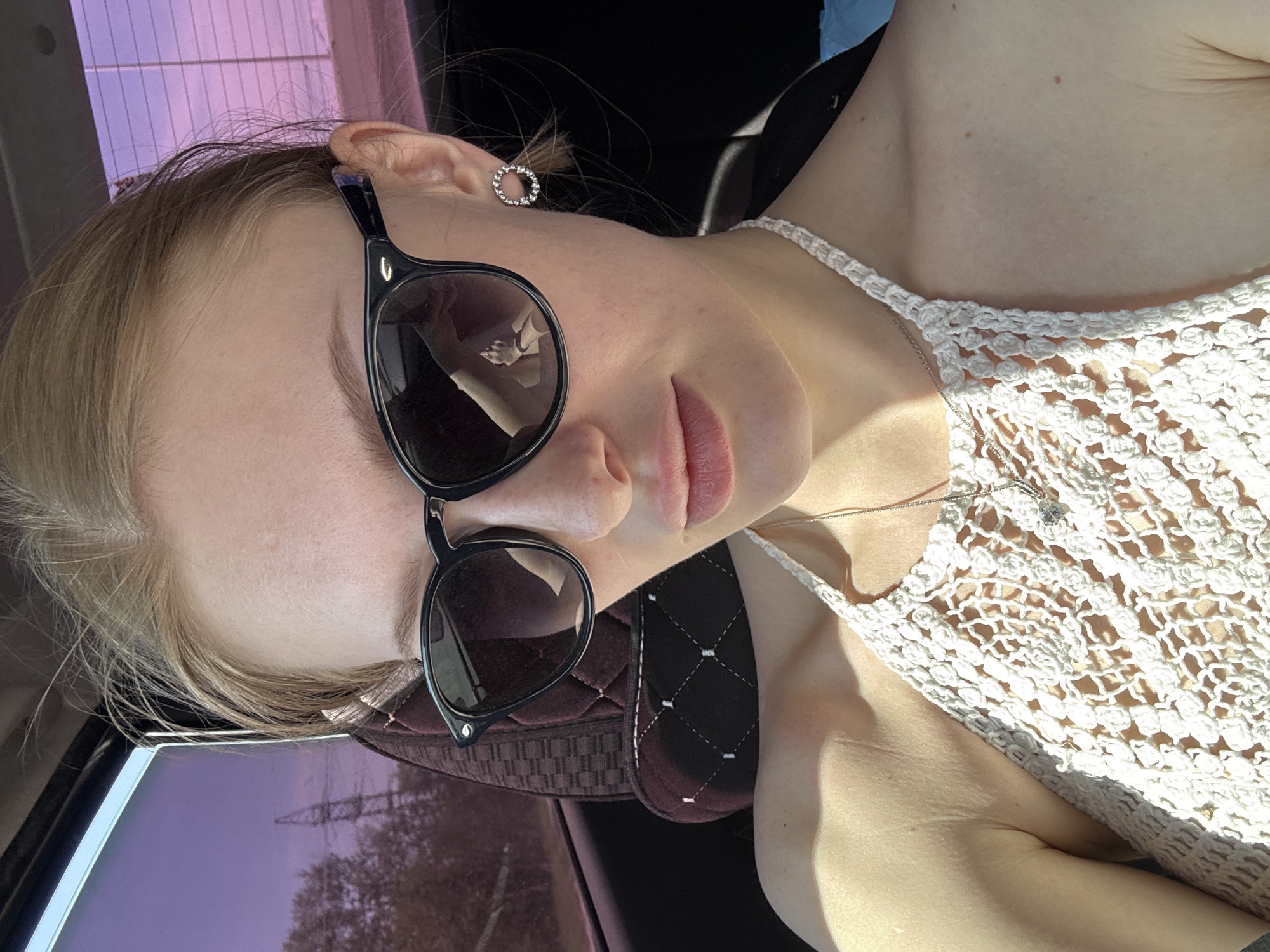 A pale-skinned person with blonde hair is wearing sunglasses and a white crocheted top. The person is sitting in a car. The location is not discernible from the image.