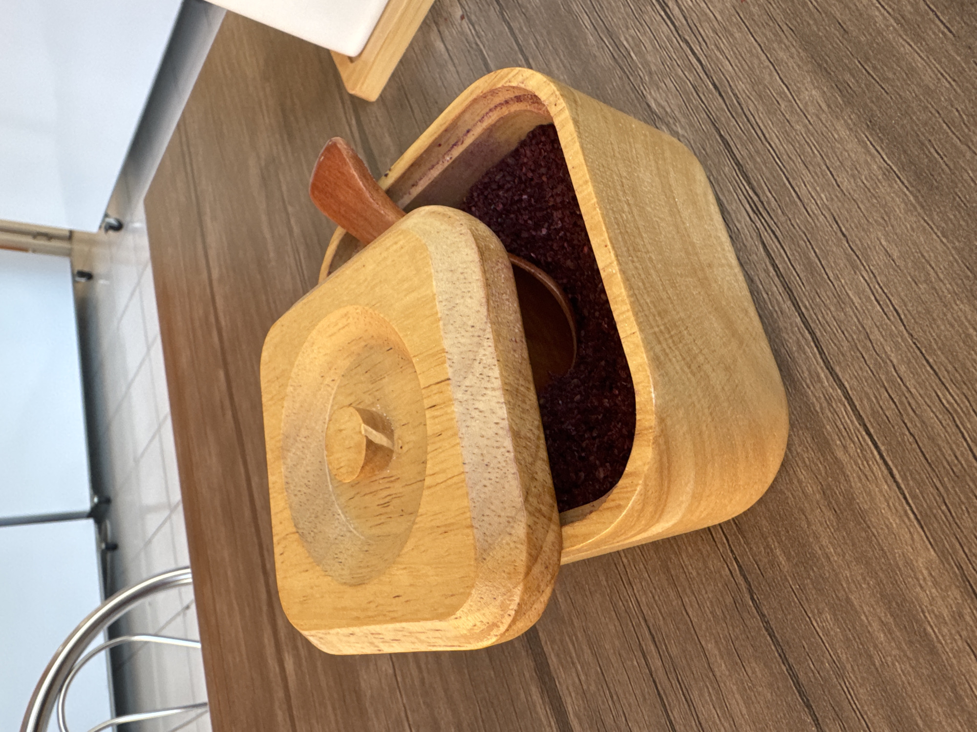An overhead shot shows a light wooden box with a lid partially covering a dark red spice, possibly sumac, inside. A small wooden spoon rests in the spice. The box sits on a brown wooden table. In the blurred background, a stainless steel railing and white cabinets are visible. This scene is likely from a restaurant in Baku, Azerbaijan, offering a view into its interior design and culinary presentation.