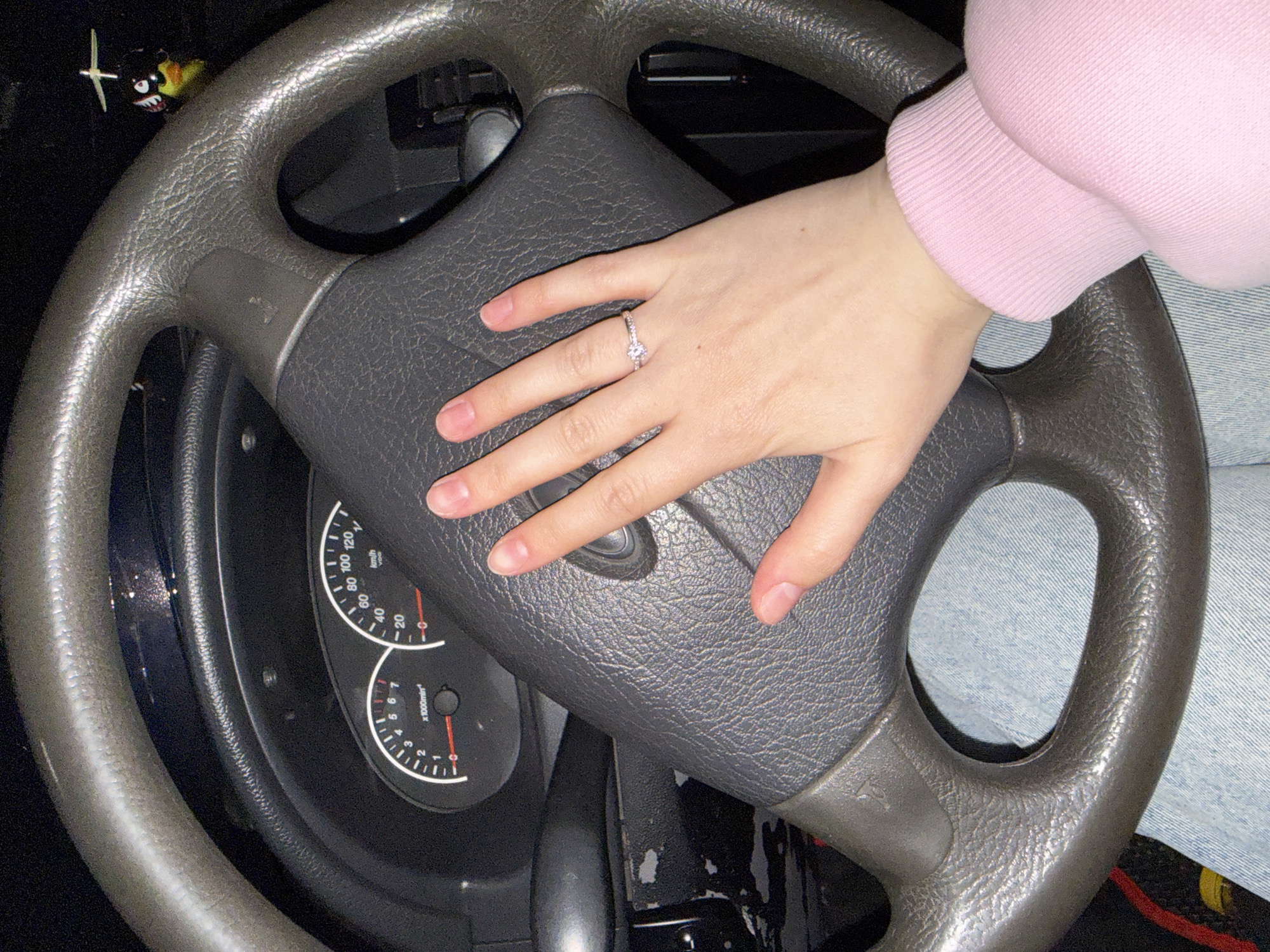 The image displays an interior view of a car, focusing on a dark gray, textured steering wheel and a person's right hand. The hand, adorned with a silver-toned ring featuring a prominent clear stone on the ring finger, rests on the steering wheel's center hub. The person wears a light pink long-sleeved top with a ribbed cuff and light blue denim jeans, partially visible on the right side of the frame.
Behind the steering wheel, sections of the instrument cluster are visible, including a speedometer marked up to 120 km/h and a tachometer indicating "x1000 min^-1" with readings up to 7. The display clearly shows "km/h", indicating a metric speed system. The overall lighting is dim, suggesting the photo was taken during evening, night, or in a dimly lit environment. A small, distinctive ornament, possibly a toy with shark teeth and eyes, is visible hanging or attached near the top-left spoke of the steering wheel. The car interior appears functional and possibly belongs to an older model. This scene is set in Zhigalki, Russia.