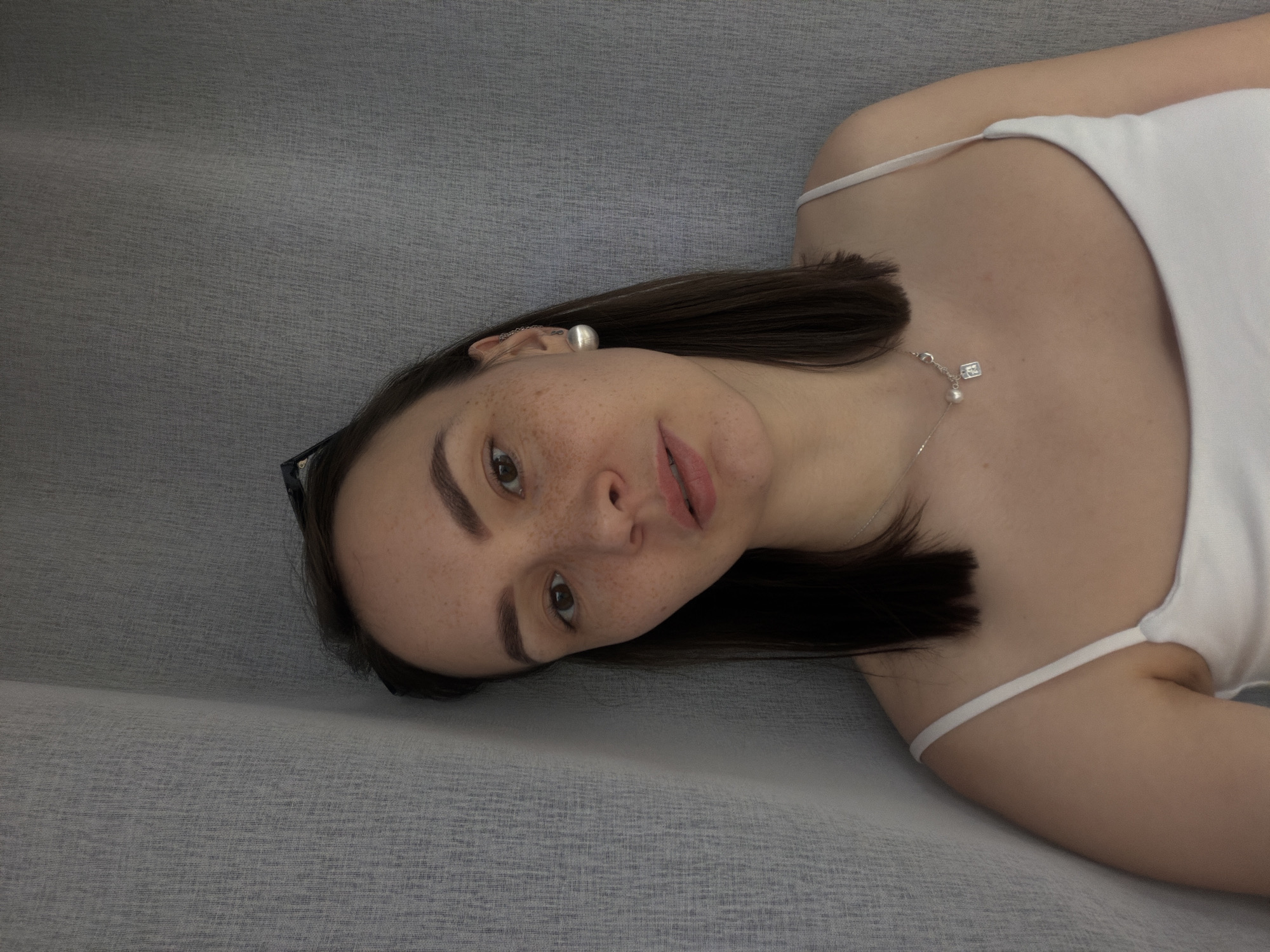 A young woman lies on a gray textured surface. She is wearing a white tank top and a silver necklace with a small pendant. She has dark brown hair, brown eyes, and freckles. She is wearing a silver earring on her left ear. A pair of glasses is visible near her hair.
The location information is not discernible from the image.