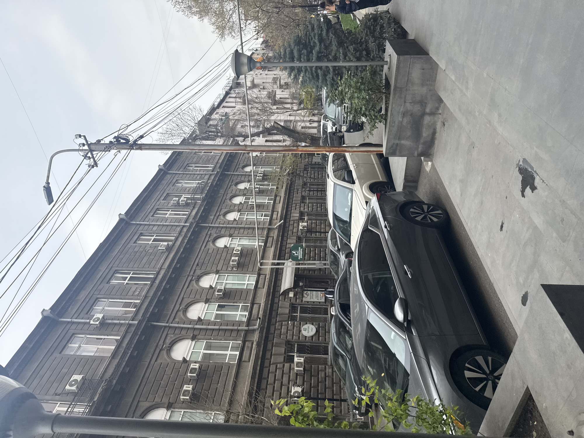 The image shows a street scene in Yerevan, Armenia, featuring a dark-gray, multi-story building with a facade of textured stone blocks and evenly spaced, arched windows. Each window has an air conditioning unit installed. The building's entrance is partially obscured by a small green sign. Parked parallel to the building are several cars in shades of black, white, and gray. A streetlight pole is visible along the edge of the sidewalk. The sidewalk is made of gray concrete slabs. In the background across the street, there's a lighter-colored building. Some trees and a person are standing on the sidewalk on the other side of the street. The sky appears overcast.