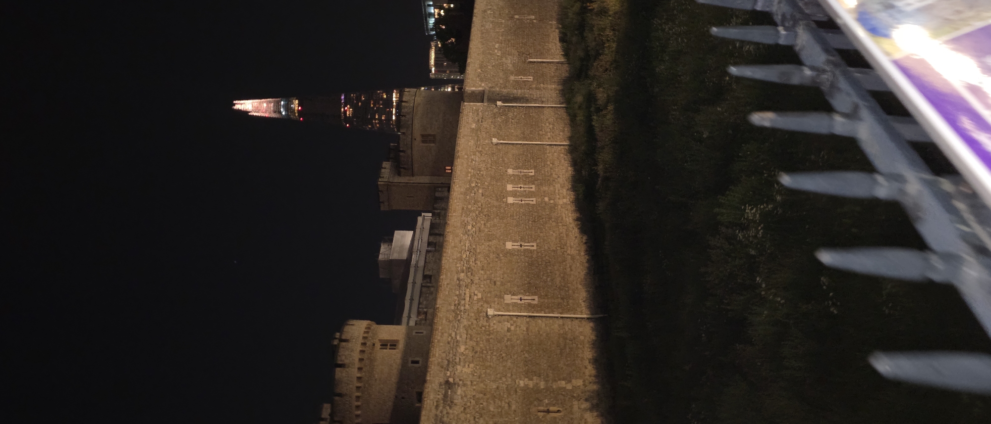 The image captures a nighttime view of the Tower of London, a historic castle on the north bank of the River Thames in central London, United Kingdom. In the foreground, a dark, grassy area is visible, separated by a spiked metal fence. Beyond the fence and grass, a section of the Tower of London's stone wall extends across the frame. The wall is illuminated, revealing its textured surface and several small, rectangular windows. To the left of the wall, a round tower with battlements is visible, also illuminated. In the background, the distinctive spire of The Shard, a contemporary skyscraper, pierces the dark night sky, its upper sections lit with red and white lights. Other buildings and city lights are faintly visible in the distance. The sky is clear and dark, with no visible stars or moon. The overall impression is one of historical architecture juxtaposed with modern urban development.