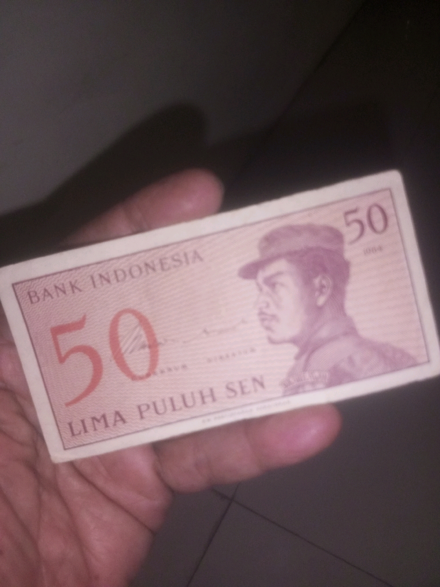 An Indonesian 50 sen banknote from 1964 is held by a person's hand. The banknote features a portrait of a man in a hat and the text "BANK INDONESIA" and "LIMA PULUH SEN". The background is dark and indistinct, suggesting an indoor or low-light environment. The specific location in Kediri City, Indonesia, cannot be determined from the image.