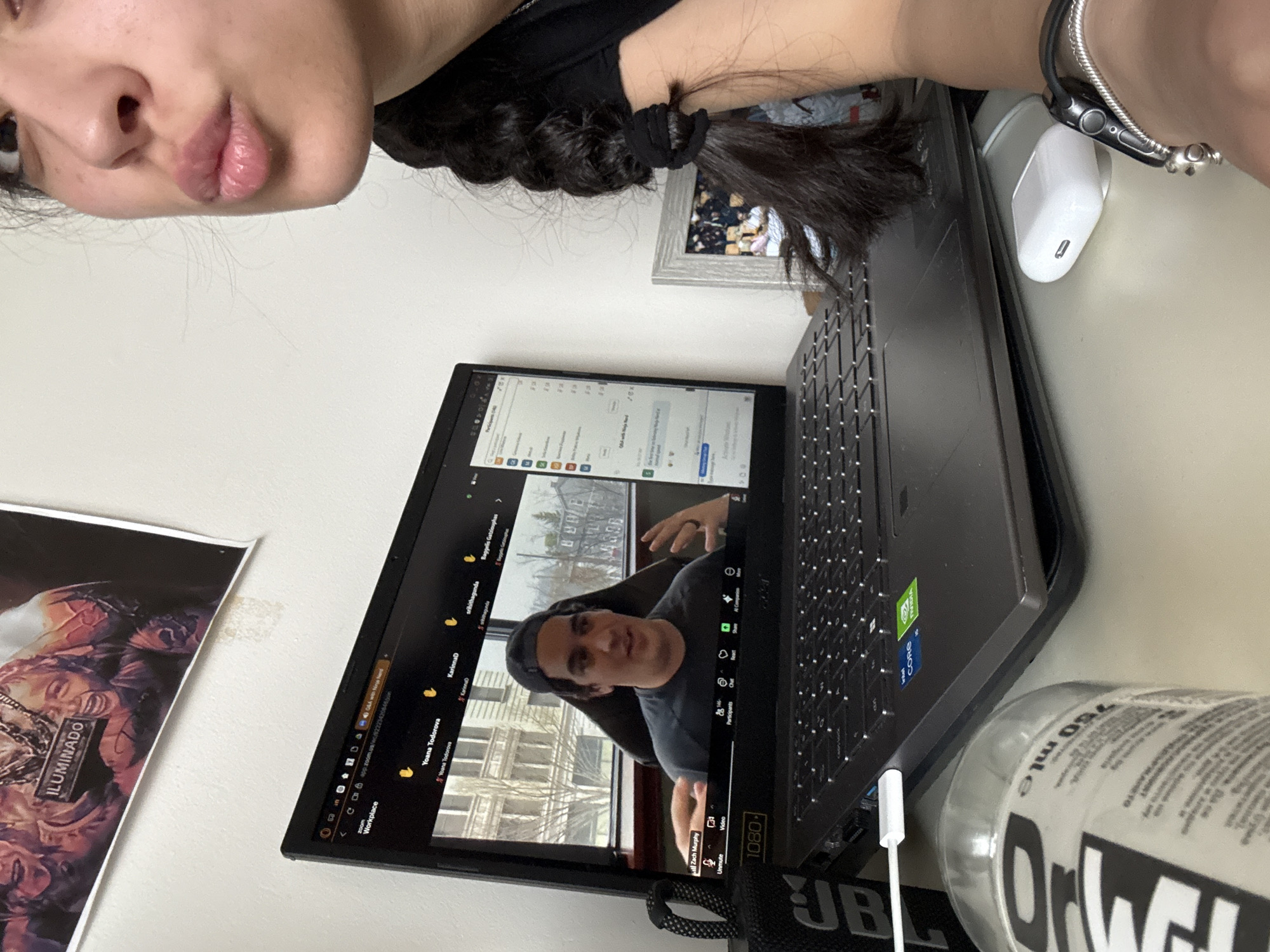 The image shows a person (possibly female) in the upper left corner, with the lower part of their face visible. They are using a laptop to attend a Zoom meeting. On the screen, another person identified as Zach Murphy is visible, possibly presenting or speaking. The Zoom interface shows participants named Yoana Todorova, KarinnaD, and srihithagunda, along with "Baggelis Gatziouphas." The laptop sits on a desk next to a water bottle labeled "Orwell." A white charger is connected to the side of the laptop. In the background, a poster or artwork is visible. A window in the Zoom background reveals a city view, but there is no reliable forensic detail that confirms it is Sofia, Bulgaria. The laptop is a dark color. A white computer mouse is next to the computer. A framed image is visible in the background.
