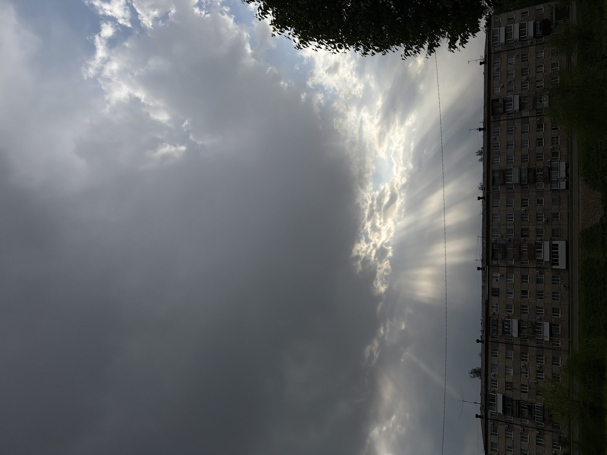 The image shows an overcast sky with sun rays breaking through the clouds, partially illuminating the side of an apartment building. The building appears to be a Soviet-era structure with multiple floors and a repetitive window pattern. Trees and other vegetation are visible along the edge of the building and in the foreground. The image suggests a late afternoon or early evening scene, with the sky appearing dramatic and partially illuminated. The exact location in Birobidzhan, Russia, cannot be determined from the image alone. No people or specific actions are visible.