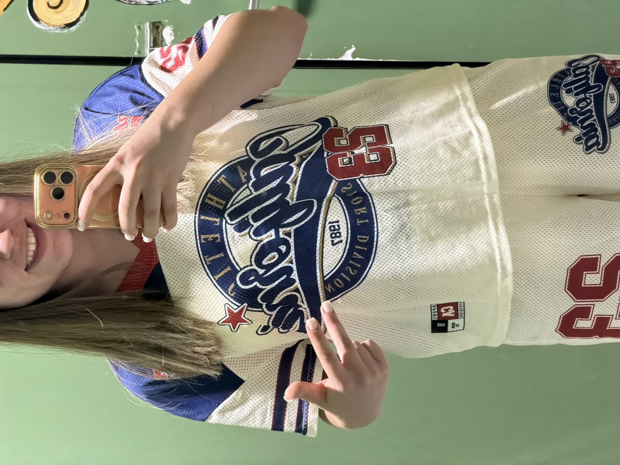 A person is holding up their cell phone to take a picture of themself. They are wearing a white mesh shirt that is adorned with red and navy blue designs. On the shirt, the word "California" is displayed in a stylized script. The number 53 can be seen in red lettering on the shirt as well. The person is posing with their right hand showing only their pointer and middle finger in front of the shirt. Behind the subject is a plain green wall. It is impossible to determine the location in the city of Arroyo de la Miel-Benalmádena Costa, Spain, without more information.
