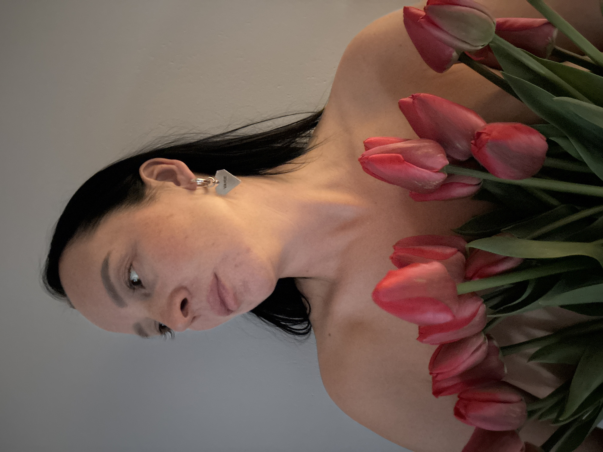 A woman with dark hair is pictured with pink tulips. She is wearing a silver earring. The background appears to be a plain, light-colored wall. No other objects or people are visible in the scene. The location, Chaikovsky, Russia, is not discernible from the image.