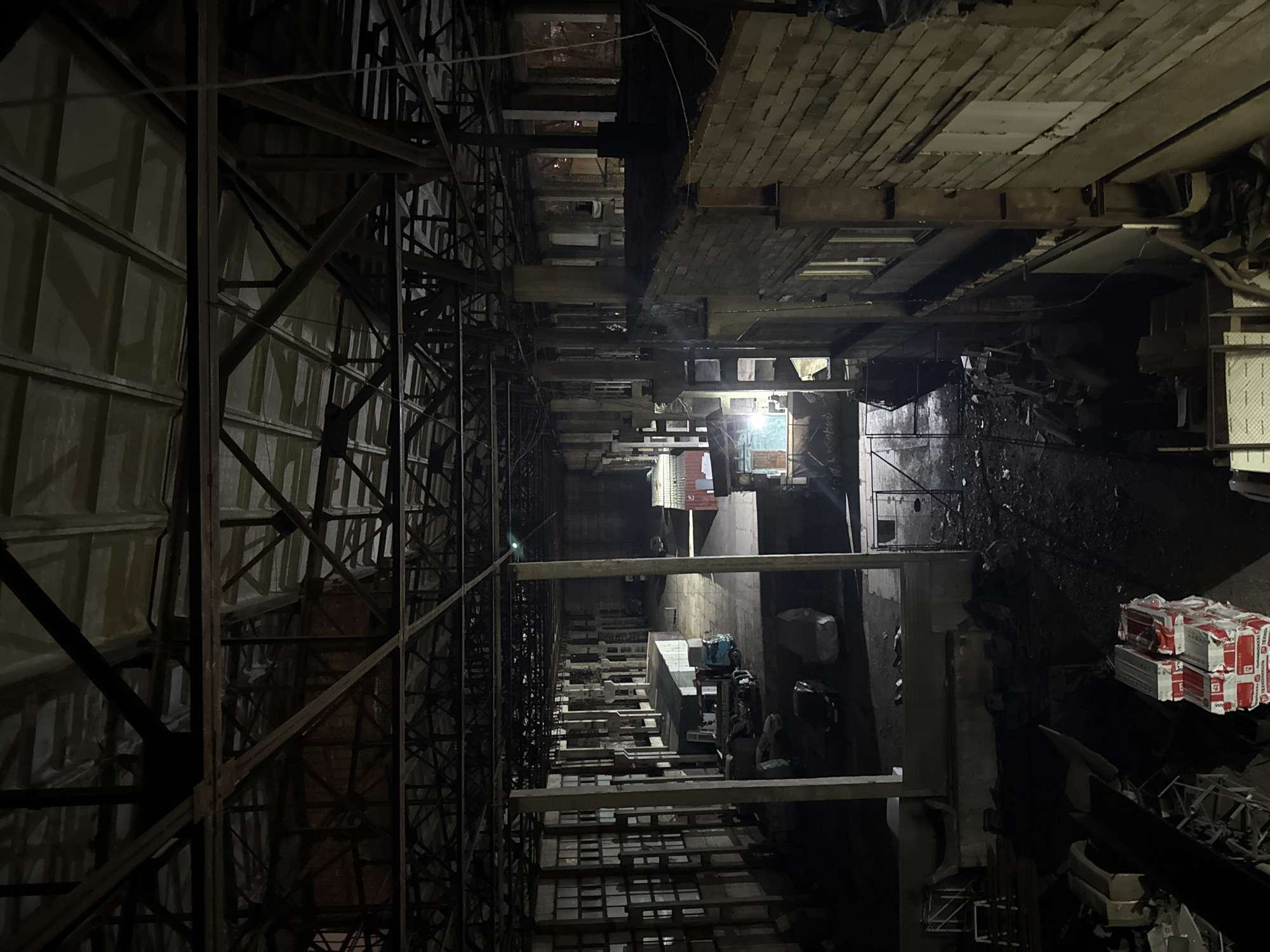 The image depicts a multi-story industrial building or complex in a state of disrepair or construction. The view is oriented vertically, looking down into the building's interior. On the left is scaffolding or structural supports. The floors of the building appear to be made of brick and concrete. Debris, equipment, and stacks of materials are scattered throughout the lower floors. There are no people visible in the image. There is a label visible on one of the stacks, reading "TERMIX" in Cyrillic text. The photo was taken in Krasnoyarsk, Russia.
