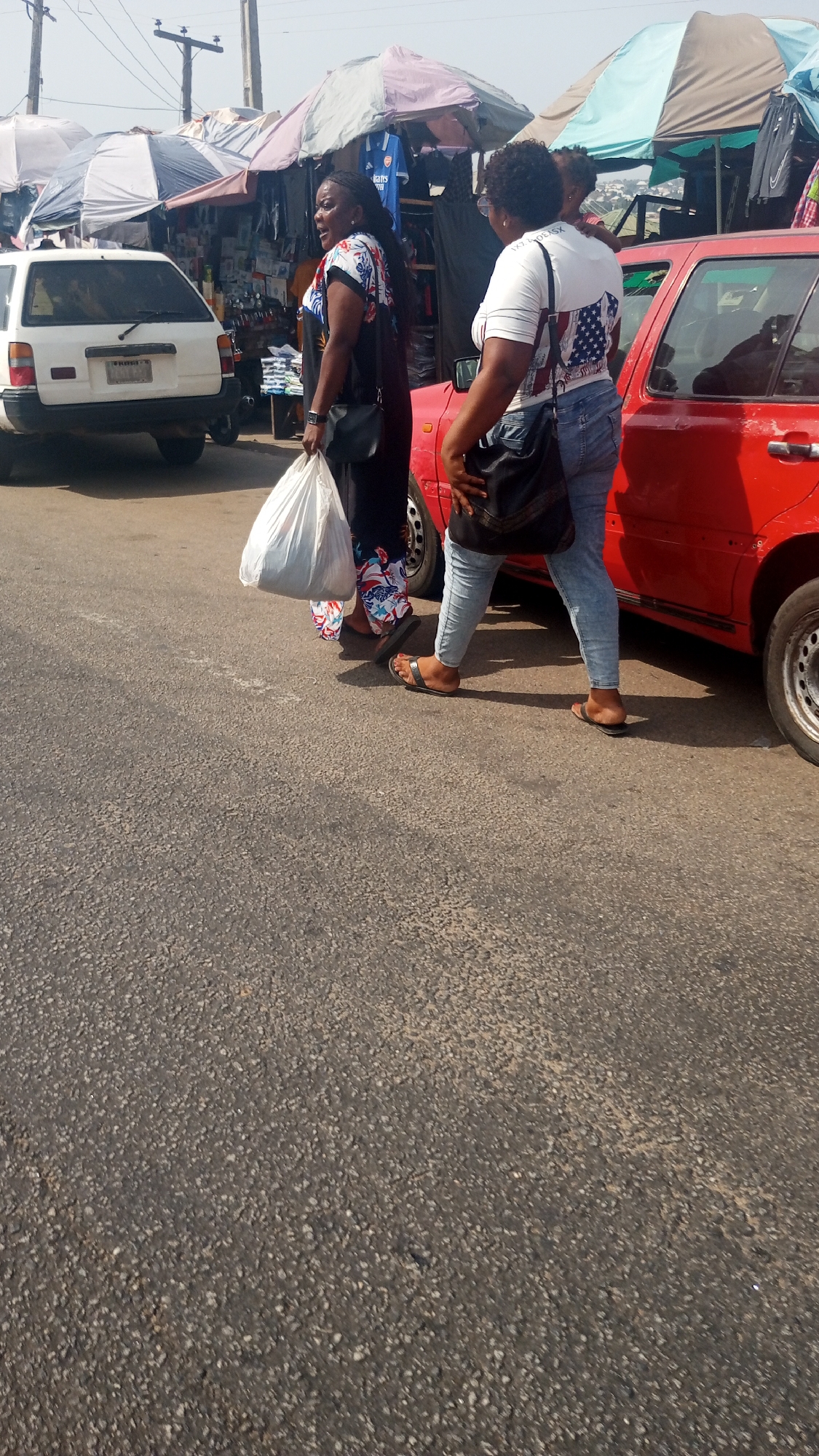The image depicts a bustling street scene in Lokoja, Nigeria, under bright daylight. In the foreground, two women walk on the right side of an asphalt road. The woman on the left, facing towards the left, wears a black top with colorful patterns, dark pants, and black sandals. She carries a large white plastic bag in her left hand and a black shoulder bag. Her mouth is open, suggesting she might be speaking or calling out. The woman on the right is seen from behind, wearing a white t-shirt featuring a design with elements resembling an American flag on the back, light blue jeans, and dark sandals. She carries a black shoulder bag.
To the left of the women, a white car, likely a sedan or station wagon, is parked with its rear visible, showing a license plate starting with "KW". To their right, a red four-door car is partially visible.
The background reveals a lively market area with numerous stalls shaded by large, colorful umbrellas. These stalls display various goods, including clothing and other items. A blue jersey with the word "ARSENAL" prominently visible, possibly along with a sponsor name like "FLY EMBIRATES", hangs in one stall. Power lines and utility poles are also visible against the clear sky. The woman in the white t-shirt has partially visible text on her back, reading "IXY COOL X". The scene captures everyday activity in a vibrant urban market setting.