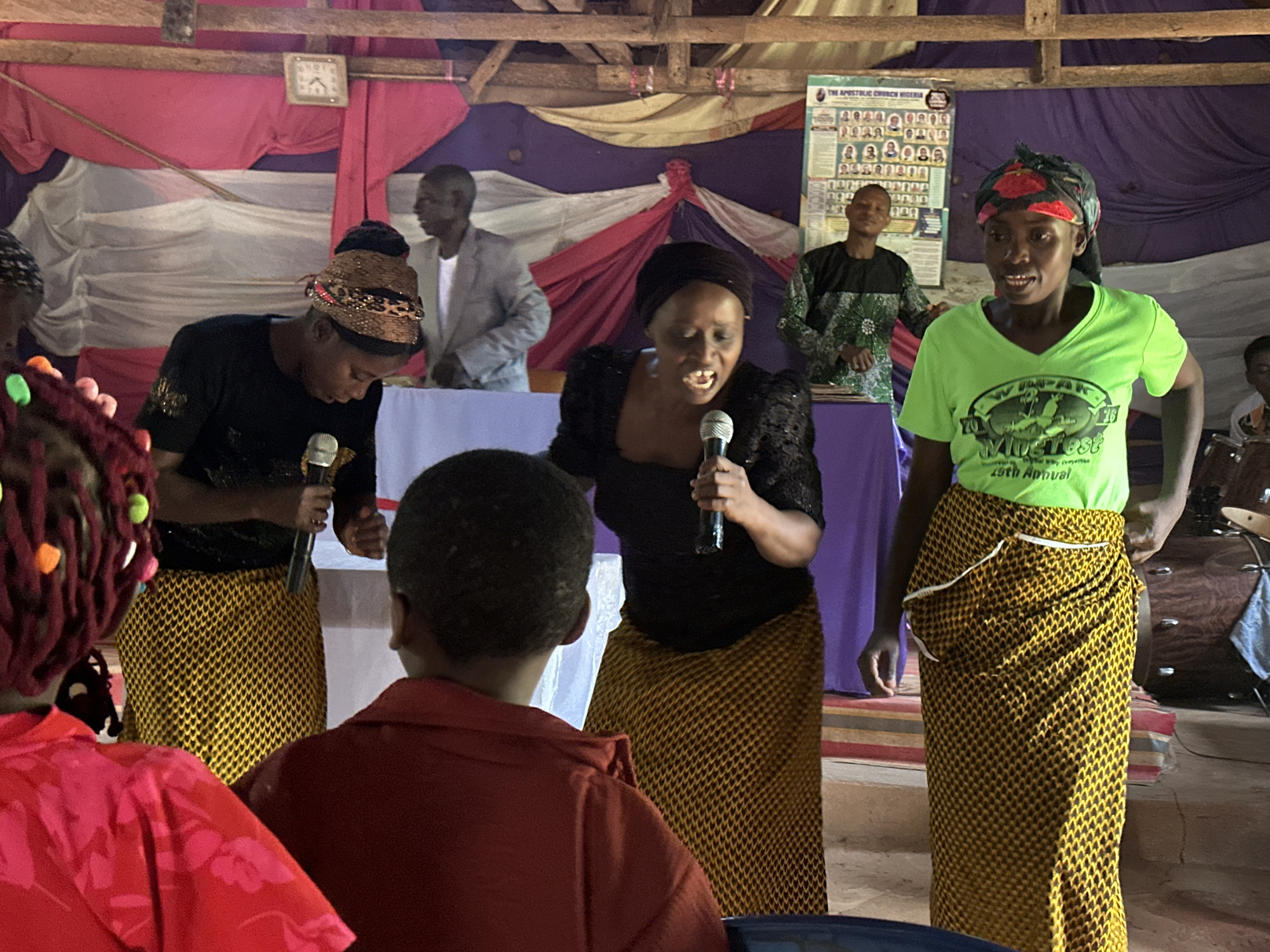 The image captures a scene inside a building, likely a church, in an unknown location within the city of Unknown, Unknown. Three women are standing near the front, each holding a microphone, suggesting they are singing or speaking. They all have on patterned yellow and black wraparound skirts. The woman on the left is in a black short-sleeved top, and she is wearing a patterned head wrap. The woman in the center is wearing a black, puffy sleeved top with a black head wrap. The woman on the right is wearing a lime green t-shirt with a graphic design and a black headwrap. Behind them, two men are standing at a table covered with a purple tablecloth, suggesting they are leading the service or event. Further back, a banner with the words "THE APOSTOLIC CHURCH NIGERIA" and multiple pictures of people on it is visible. In the foreground, there are members of the audience. There is a clock on the wall. The backdrop features draped fabric in white, red, and purple. The overall atmosphere suggests a church service.