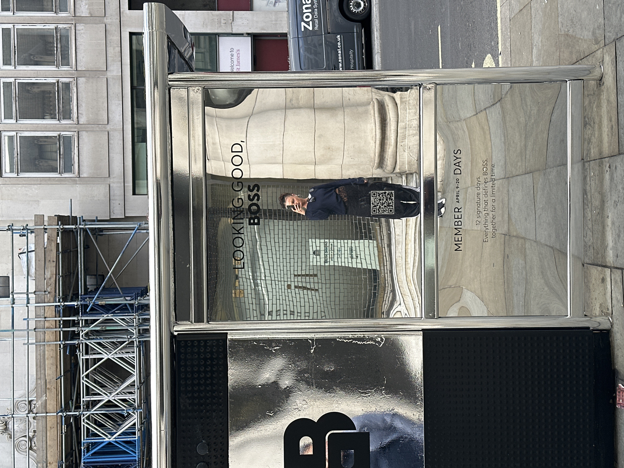 The image depicts a reflective advertising panel for BOSS, situated on a sidewalk in London, United Kingdom. The advertisement features the text "LOOKING GOOD, BOSS," accompanied by an image of a man using a phone, and the text "MEMBER APRIL 9-20 DAYS," promoting a limited-time event. A QR code is visible on the panel. Reflections in the panel show surrounding buildings, a construction scaffolding, and a parked van with "Zonal Retail Data System" signage. The sidewalk is composed of stone. The bottom portion of the advertising panel displays the BOSS brand logo in a reflective silver material against a black background.