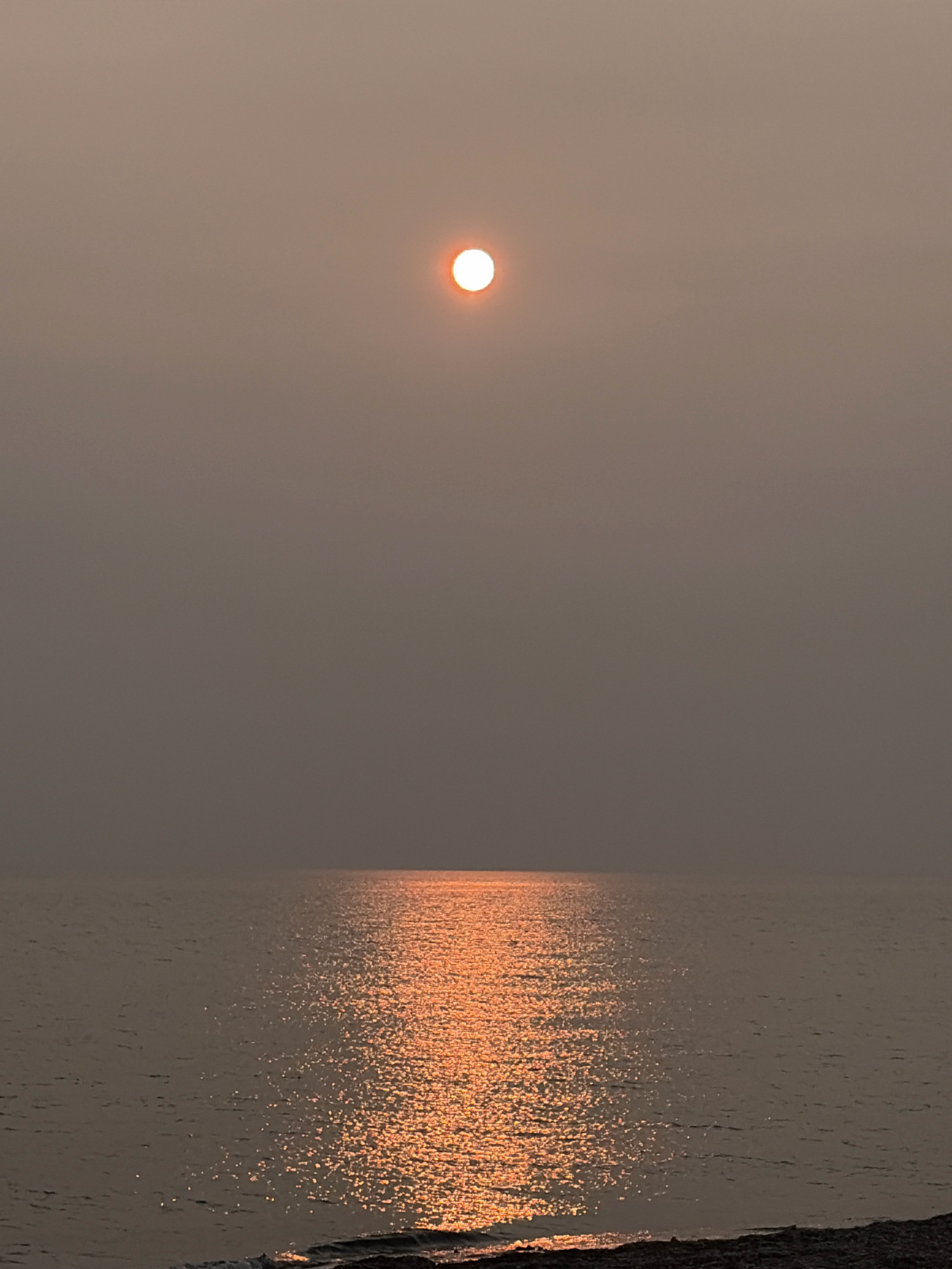 The image shows a hazy sunset over a body of water, likely Lake Ozerskoe in Ozerskoe, Russia. The sun, a bright orange orb, is positioned in the upper center of the frame, partially obscured by a thick haze. A shimmering reflection of the sun extends across the water's surface, creating a path of golden light. The water itself appears calm, with gentle ripples. The foreground shows a dark, rocky shoreline. The overall atmosphere is subdued and muted due to the atmospheric conditions. No people or animals are visible in the scene.