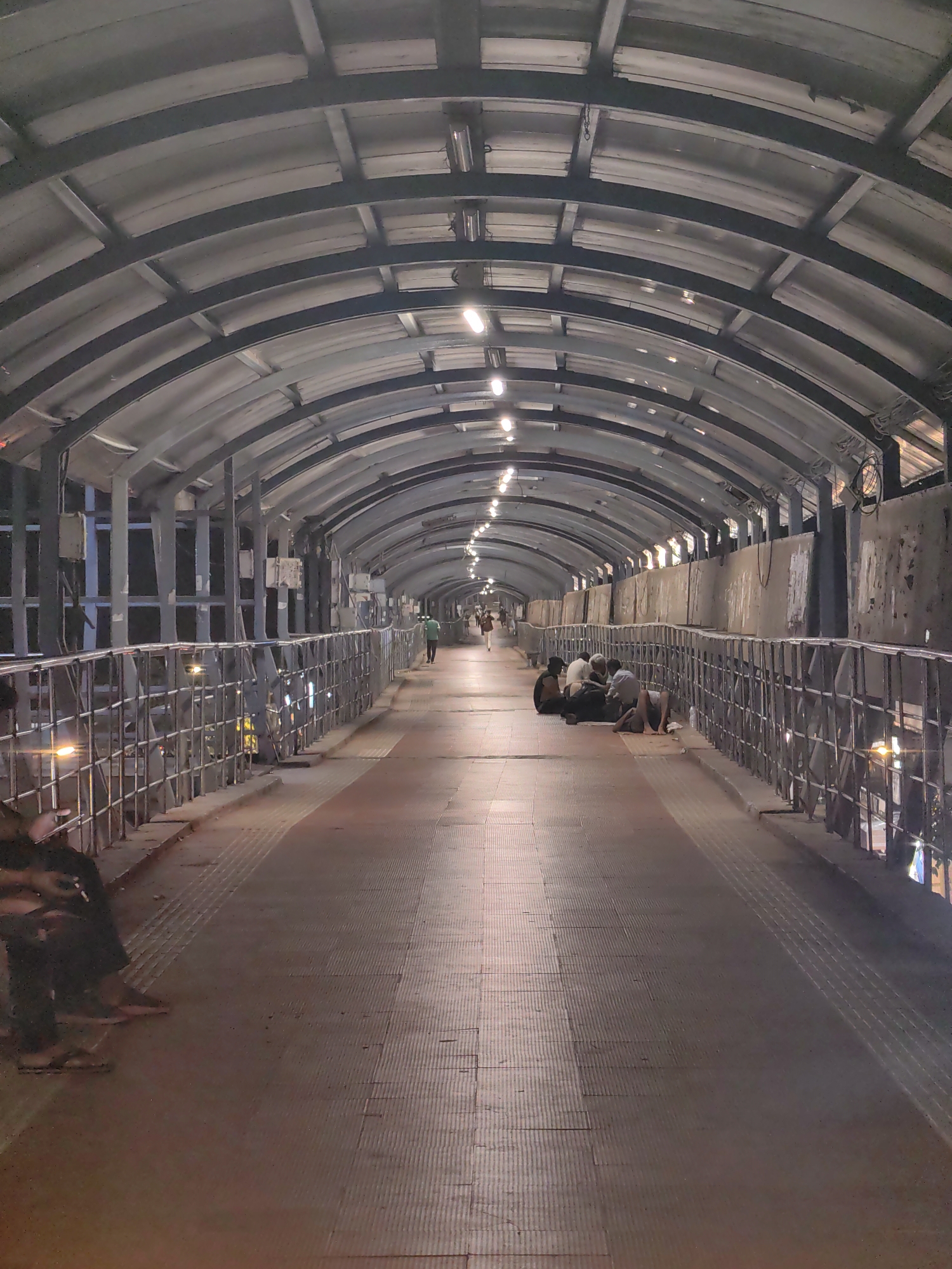 This is a vertical, eye-level, medium-shot photograph taken at night on a covered pedestrian walkway, likely in Mumbai, India. The walkway is enclosed by a corrugated metal roof supported by a dark metal frame, with rows of fluorescent lights illuminating the path. The walkway itself is paved with textured red tiles and has a metal railing on both sides.
On the right side of the walkway, a group of at least five individuals are seated on the ground, appearing to be resting or sleeping. They are dressed in casual clothing. Further down the walkway, a single person in a green shirt is walking away from the camera. On the left side, near the foreground, a person is seated on the ground with their legs extended.
The environment suggests a public space, possibly a footbridge connecting different parts of a transportation hub or a busy area, given the lighting and the presence of people. The overall impression is one of urban infrastructure at night, with a mix of transient occupants and passersby.