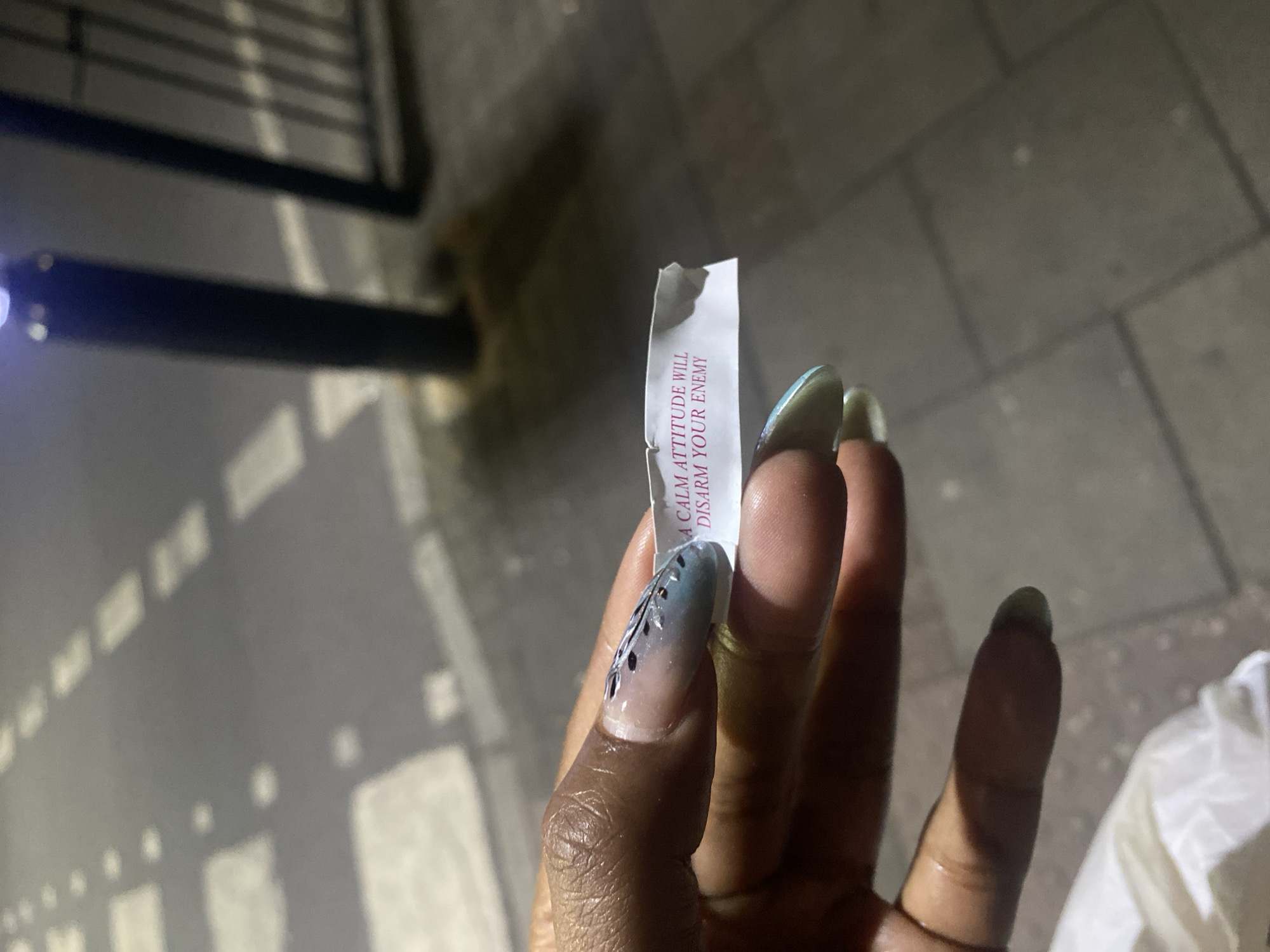 A close-up shot shows a person's hand holding a torn fortune cookie paper. The paper reads: "A calm attitude will disarm your enemy." The person has a dark skin tone and long, manicured nails with a French tip design and an additional design resembling dark feathers on one nail. The background suggests an urban setting. There is a metal railing and what appears to be a patterned pavement. The location is London, United Kingdom.