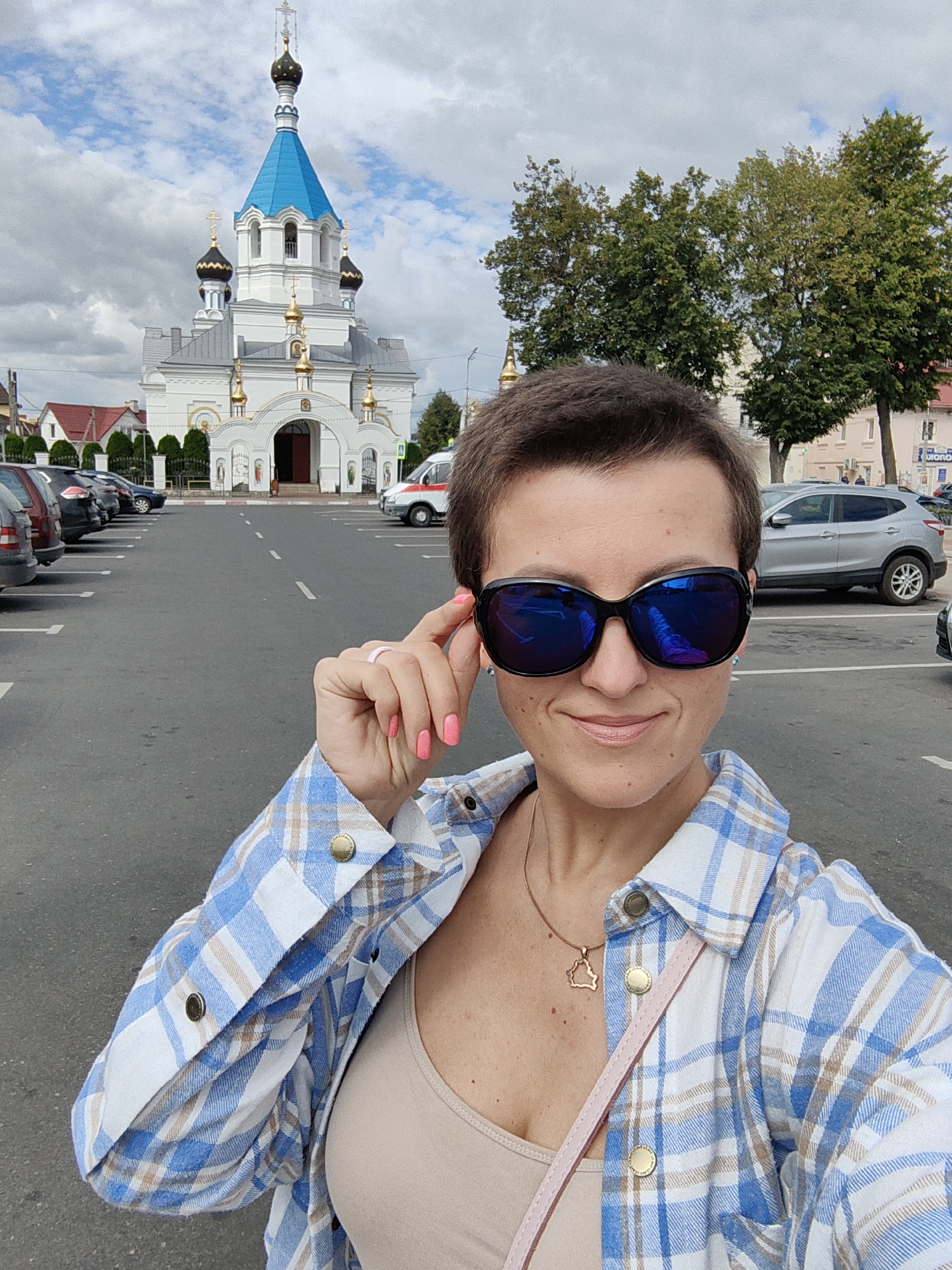 The image captures a woman posing for a selfie in a parking lot. She is wearing sunglasses, a tan tank top, and a blue and white plaid shirt. A pink purse strap is visible over her shoulder. In the background, a white and blue domed church stands prominently. To the left of the church, there are several parked cars, and to the right, trees and buildings are visible. The scene is set in the city of Pastavy, Belarus, with the church likely being the Church of the Holy Trinity, a landmark in the area. The sky is overcast with clouds.