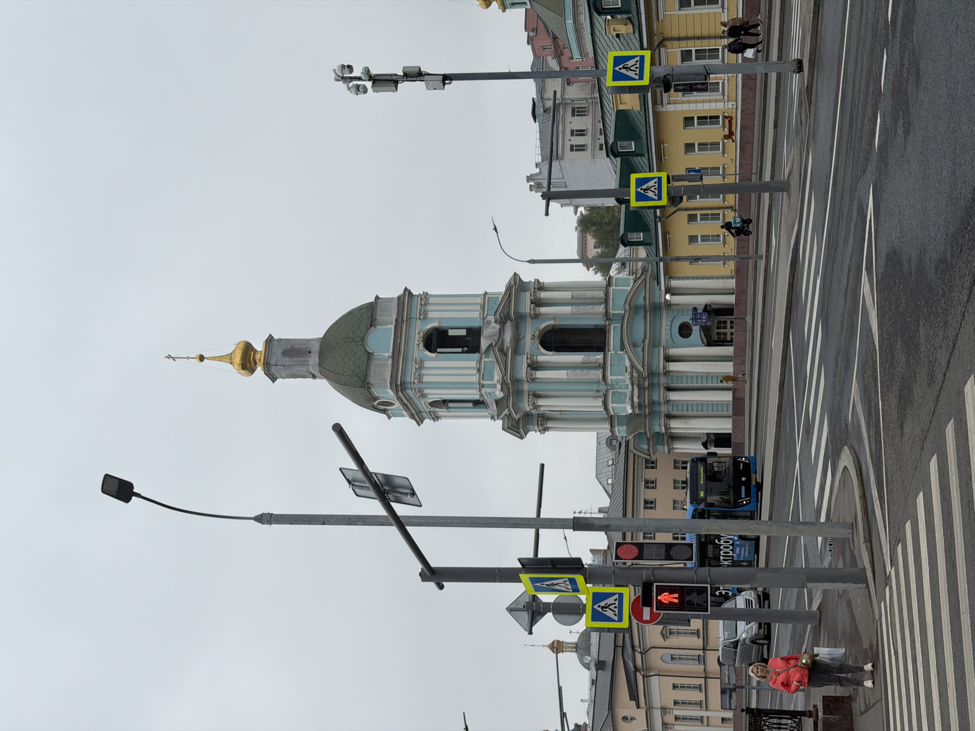 The image depicts a city street scene with a prominent architectural structure in the background, possibly a church or cathedral, characterized by its blue and white exterior and a golden dome. The sky is overcast, suggesting a cloudy day.
In the foreground, there are pedestrian crosswalks with white stripes on a gray asphalt road. Several traffic signals and streetlights are present, indicating an urban setting. Pedestrian crossing signs are visible on poles near the crosswalk.
A few people are observed in the scene. One individual, wearing a red jacket, stands on the sidewalk near the edge of the crosswalk. There are others walking on the far side of the road.
A blue bus is seen on the road, indicating public transportation. Other buildings line the street, displaying a mix of architectural styles and colors.
The street appears wet, possibly from recent rain.
