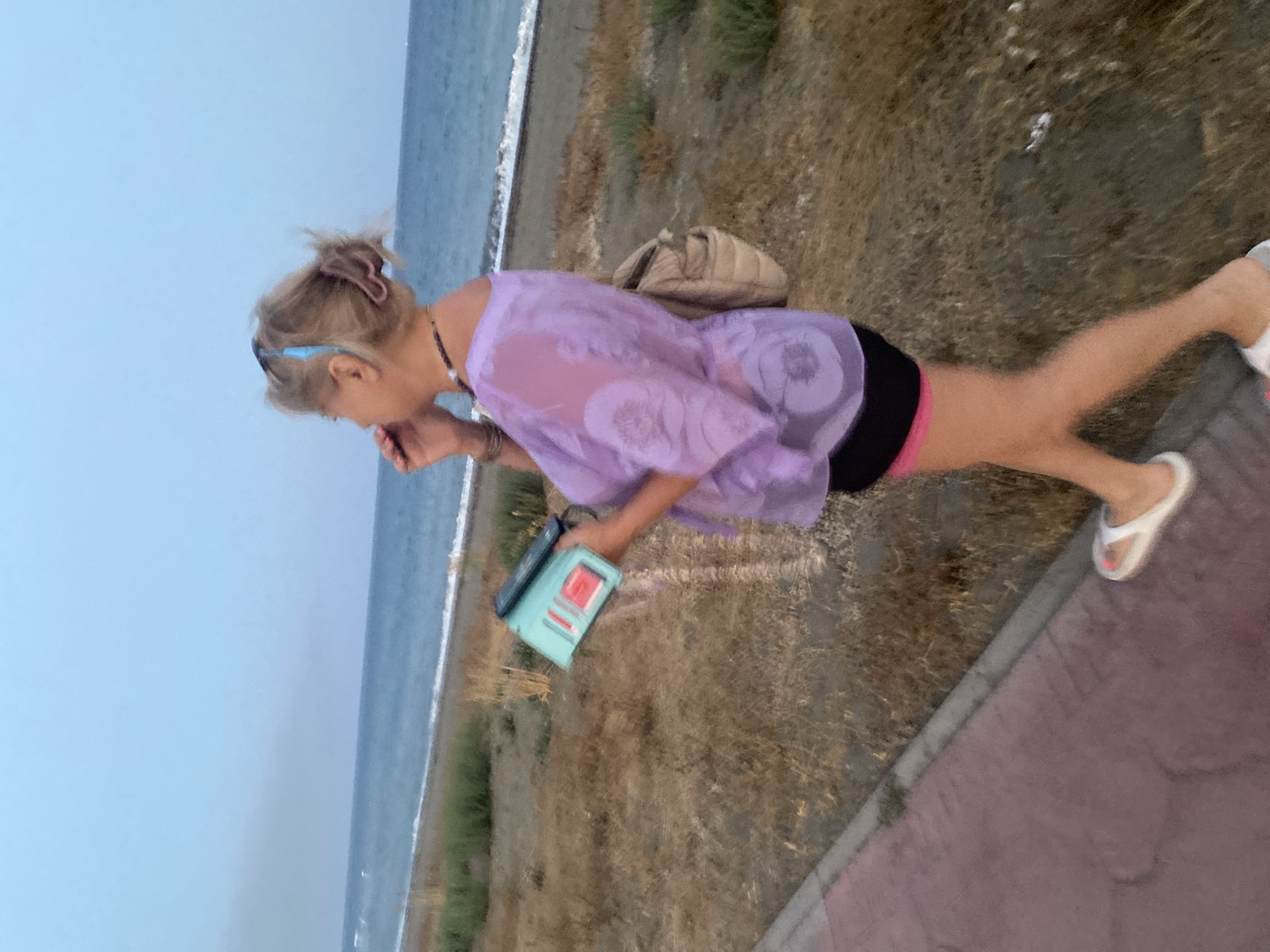 An adult female walks along a paved path beside a sandy beach with a calm sea and a pale blue sky. The woman is wearing a sheer, lavender, short-sleeved top with a floral pattern, black shorts with pink trim, and white sandals. She has blonde hair pulled back with a clip and is holding a turquoise tablet in her left hand while speaking on a dark-colored mobile phone to her right ear. A beige crossbody bag is slung over her right shoulder. The ground on the right of the path is covered in dry, sandy vegetation. The scene is likely the promenade area of Estepona, Spain, given the characteristic paving and proximity to the sea.