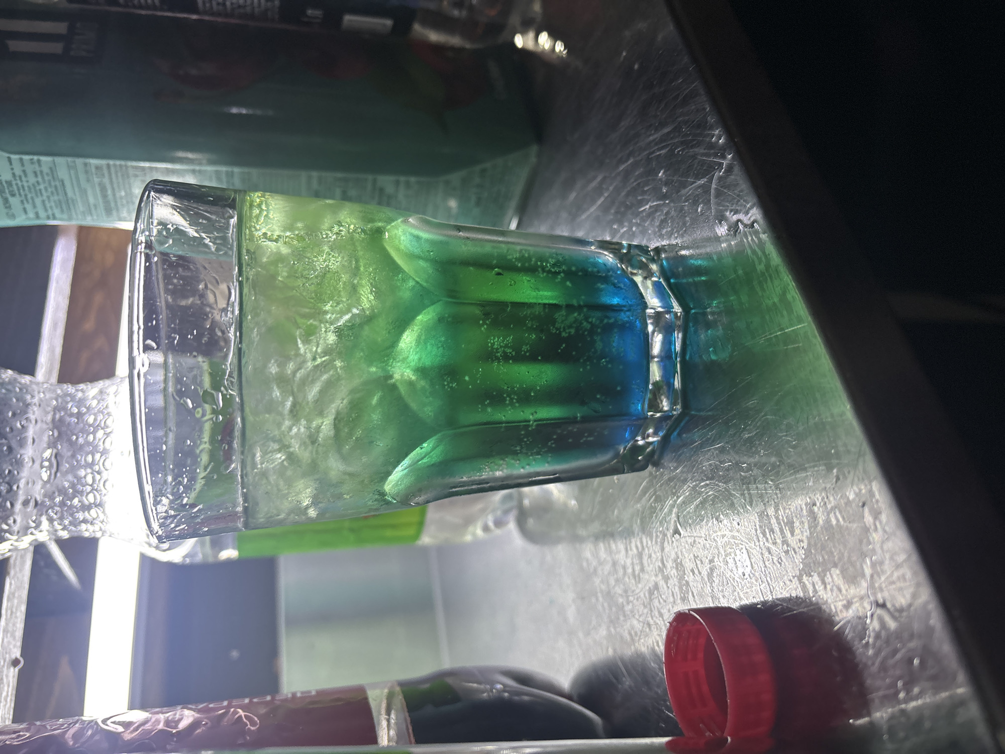 A glass of a layered blue and green beverage with ice cubes sits on a scratched metal surface. To the left of the glass is a red bottle cap. In the background, out of focus, are several bottles, one of which is green. The lighting in the scene is bright and appears to be from above. The image is a close-up shot, focusing on the drink and its immediate surroundings. There is no visual information within the image that would allow for the determination of its location in Birobidzhan, Russia, or any other specific city. The scene depicts a still life arrangement of a drink and related items on a countertop.