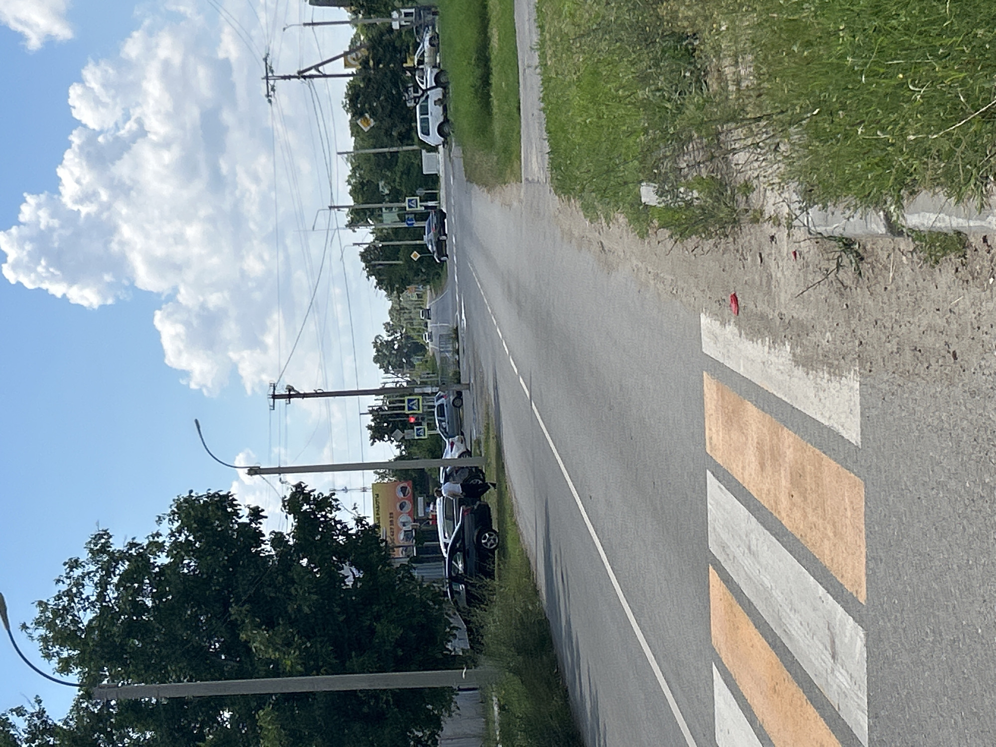 The image depicts a rural roadside scene in Dondukovskaya, Russia, on a clear, sunny day. The perspective is from the side of a paved road, with a grassy embankment to the right. Several cars are parked along the side of the road, interspersed with trees and utility poles. A pedestrian crossing with diagonal white and orange stripes is visible on the asphalt in the foreground, indicating a road crossing.
In the distance, more cars and roadside infrastructure, including traffic lights and street lamps, are visible. The sky is bright blue with scattered white, puffy clouds, suggesting it is daytime, likely late morning or early afternoon. The presence of lush green grass and trees indicates a warm season, possibly summer or late spring.
There are no distinct human activities happening, with the focus being on the parked vehicles and the general environment. One car in the middle distance has a person leaning into it, but their activity is unclear. The overall atmosphere is one of stillness and a typical day in a less urbanized area. Some faint text is visible on a yellow sign in the distance, but it is illegible.