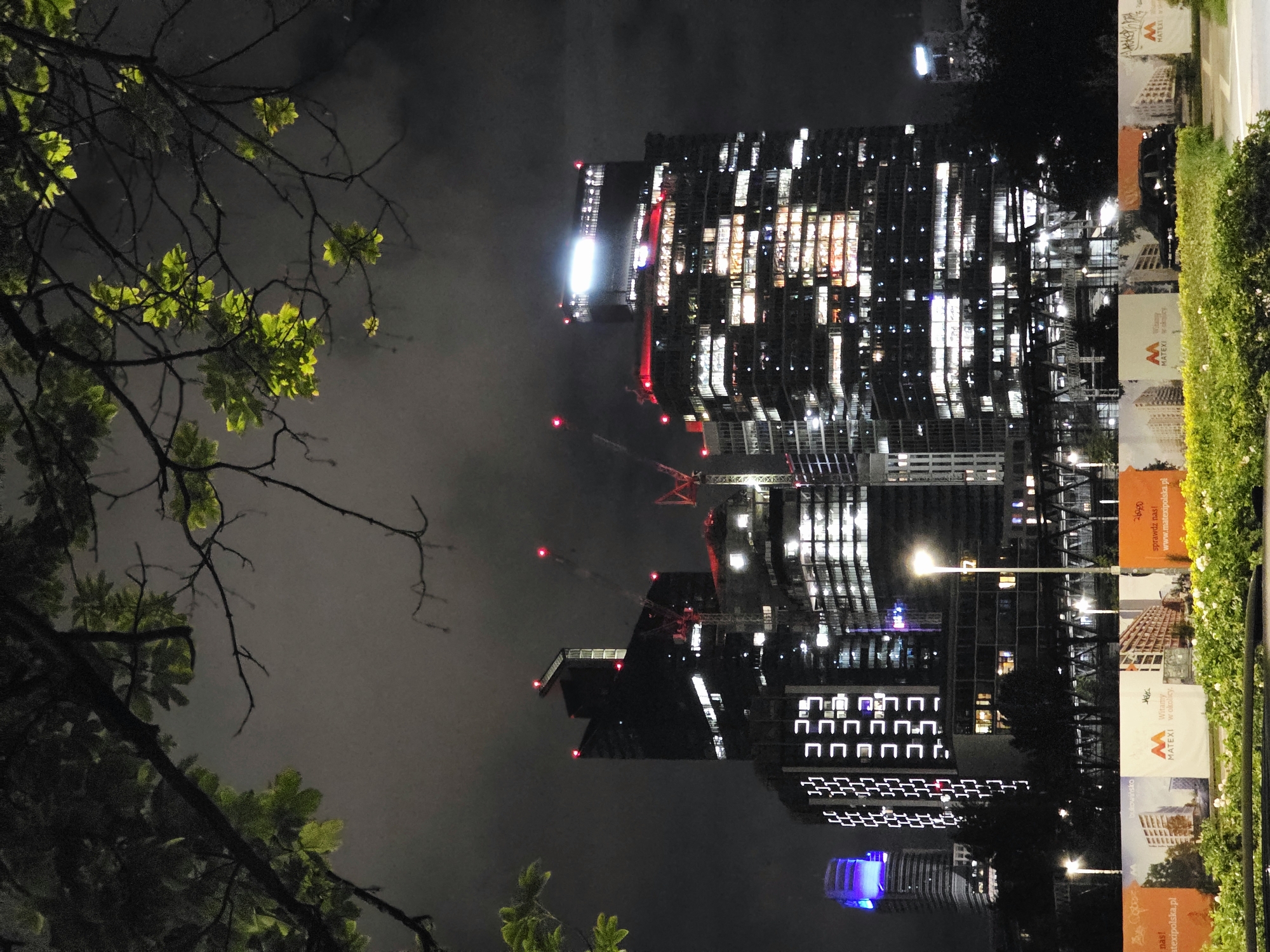 The image depicts the night cityscape of Warsaw, Poland, focusing on a cluster of modern high-rise buildings, some of which are under construction. The foreground features a leafy tree branch with illuminated green leaves, creating a natural frame for the urban scene. In the midground, several skyscrapers dominate the view. One prominent building is dark, with numerous illuminated windows revealing interior activity and a sharp, angular roof topped with red lights. Adjacent to it, another skyscraper is brightly lit, showcasing a complex facade of windows and a luminous, elongated structure at its summit. Cranes, indicative of ongoing construction, are visible among the buildings, some marked with red lights. To the left, a distinct tower with a blue illuminated cap stands out. The lower portion of the image displays a row of billboards advertising real estate developments, featuring images of residential buildings and company logos. A dark car is parked behind the billboards. Streetlights illuminate the area in front of the buildings. The overall atmosphere is urban and contemporary, set against a dark, cloudy night sky.