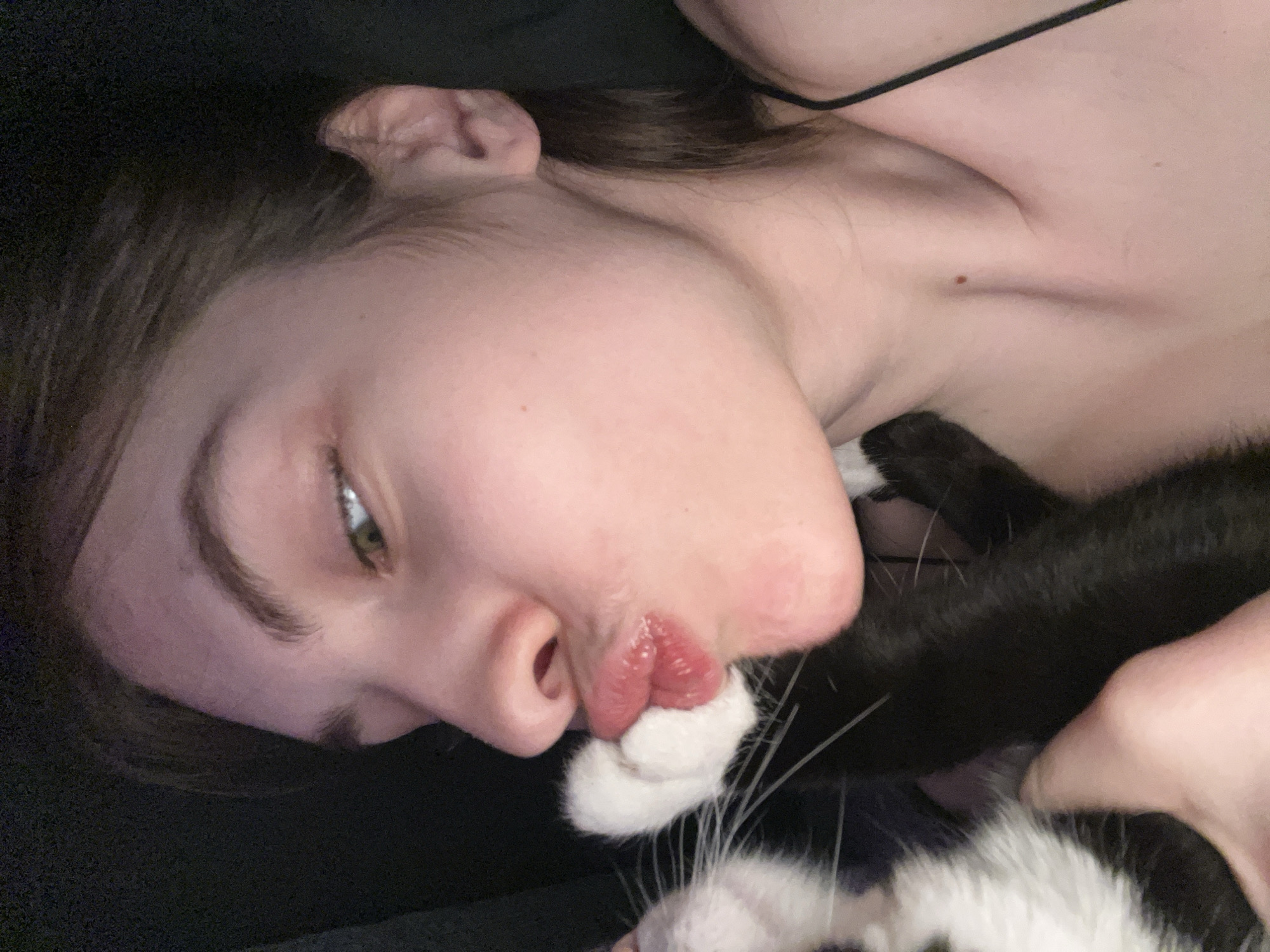 A close-up, slightly angled, indoor shot shows a person lying down next to a black and white cat. The person's face is prominent, with their lips close to the cat's paw. The cat's whiskers are visible, along with the person's eye and eyebrow. A dark-colored garment or surface is in the background. The person has fair skin and dark hair. There is no indication of the location being Saint Petersburg, Russia.