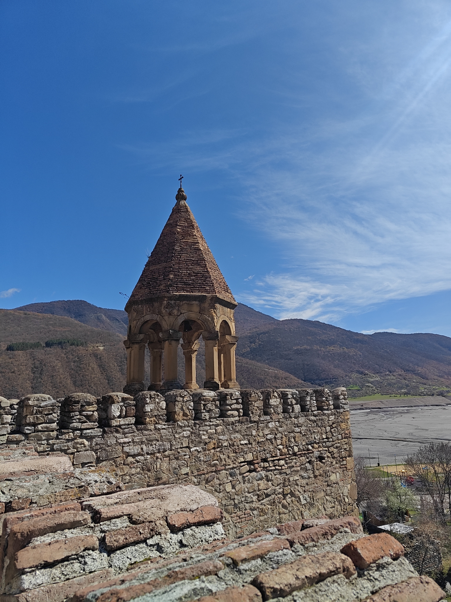 A wide-angle, eye-level view captures a historic stone bell tower and fortified walls in Ananuri, Georgia, under a bright blue sky. The bell tower, centrally positioned and slightly left of frame, features a reddish-brown conical tiled roof topped with a small cross. Below the roof, an open belfry with arches and stone columns is visible.
In the foreground, rustic reddish-brown roof tiles or brickwork are angled, leading to a sturdy stone wall with crenellated battlements extending across the midground. The wall's texture is composed of varied stones.
Beyond the fortifications, the scene opens to a vast mountainous landscape, with hillsides showing sparse vegetation in shades of brown, indicative of early spring or late autumn. A wide valley floor is visible to the right, appearing as a dry riverbed or a low reservoir level, with some green patches and distant trees. The sky is a vivid blue, with delicate, wispy white clouds. Bright sunlight illuminates the entire scene, with a subtle lens flare in the upper right.
No people or specific activities are depicted. There is no visible text in the image. The media conveys a sense of enduring history and scenic natural beauty.