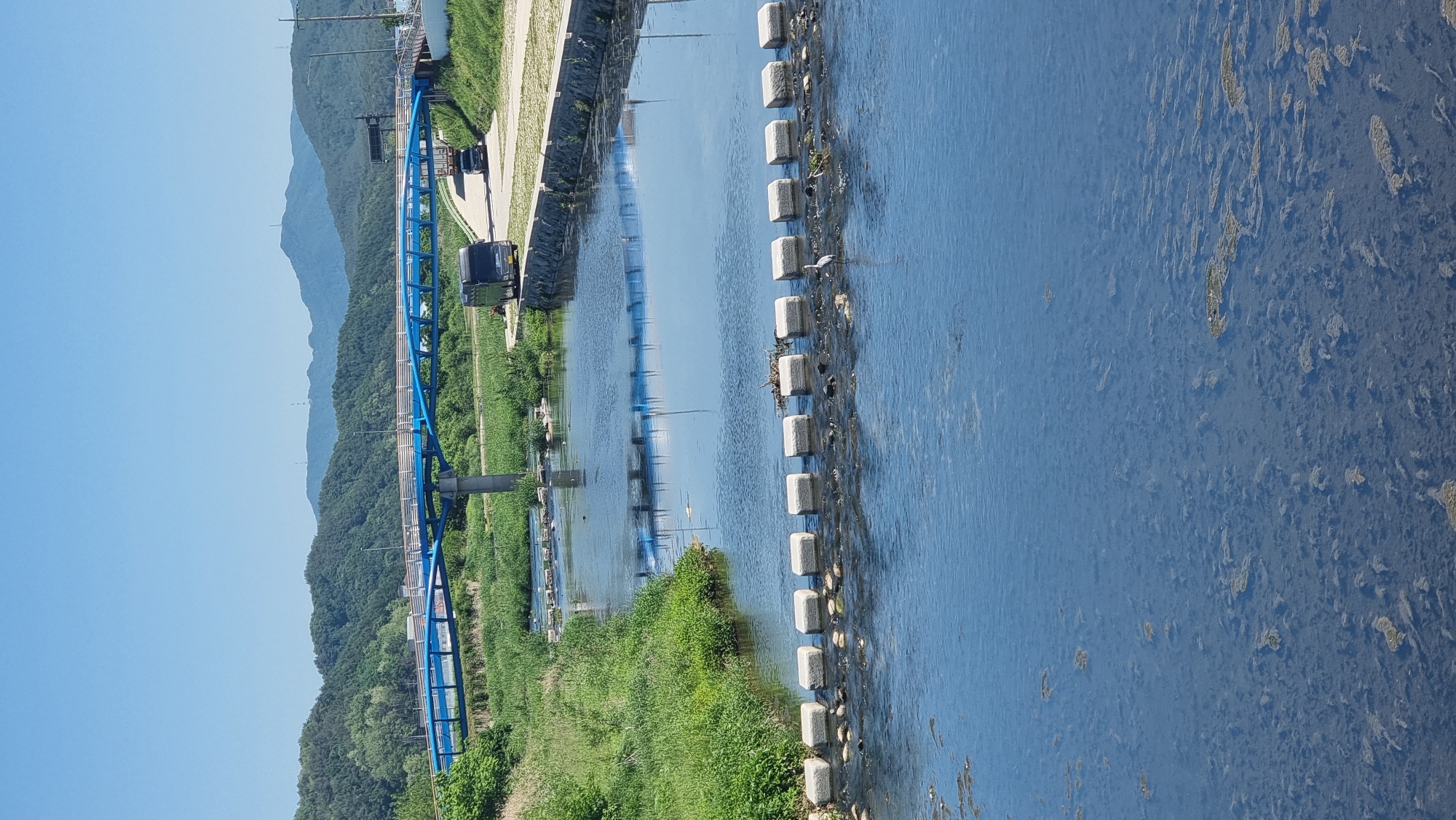 The photograph depicts a river flowing through the urban region of Dunnae-myeon, South Korea. A blue metal bridge extends over a portion of the river and adjacent greenery. The water surface has concrete stepping stones across part of the river and is reflective. Vegetation flanks both banks of the water body. In the background, a mountain range ascends into a clear sky. A car is present on the river's edge, next to the river's edge.