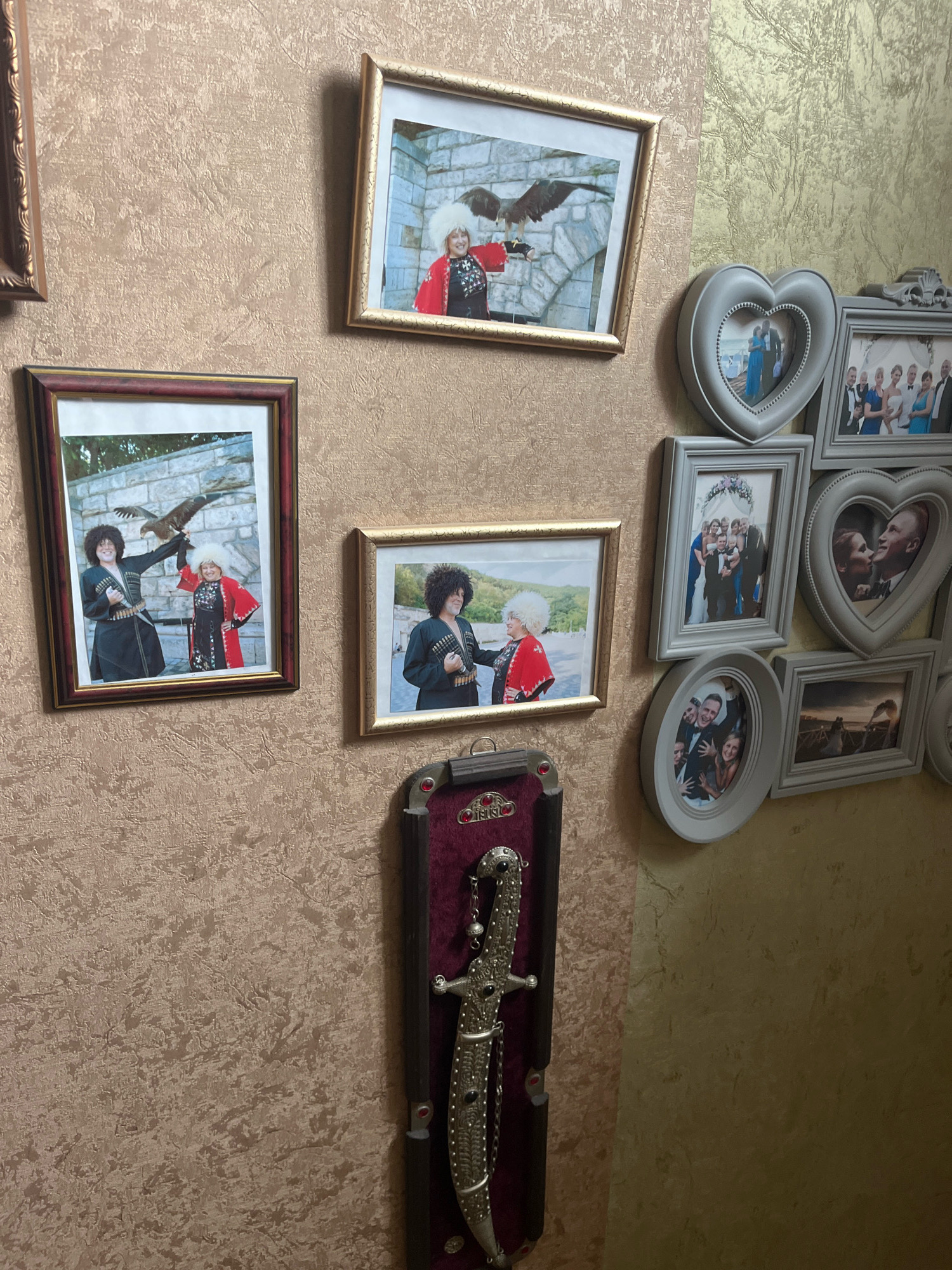 In a room with textured wallpaper, a collection of framed photographs and a decorative dagger are displayed on the wall. One framed photograph shows two individuals dressed in traditional attire, one holding a bird of prey. Another photograph depicts a group of people in formal wear, possibly a wedding party. Other framed pictures include couples and group portraits. A decorative dagger with an ornate handle and sheath is mounted on the wall. The overall impression is of a personal display of memories and cultural artifacts. The location is specified as Vladimirovka, Russia.