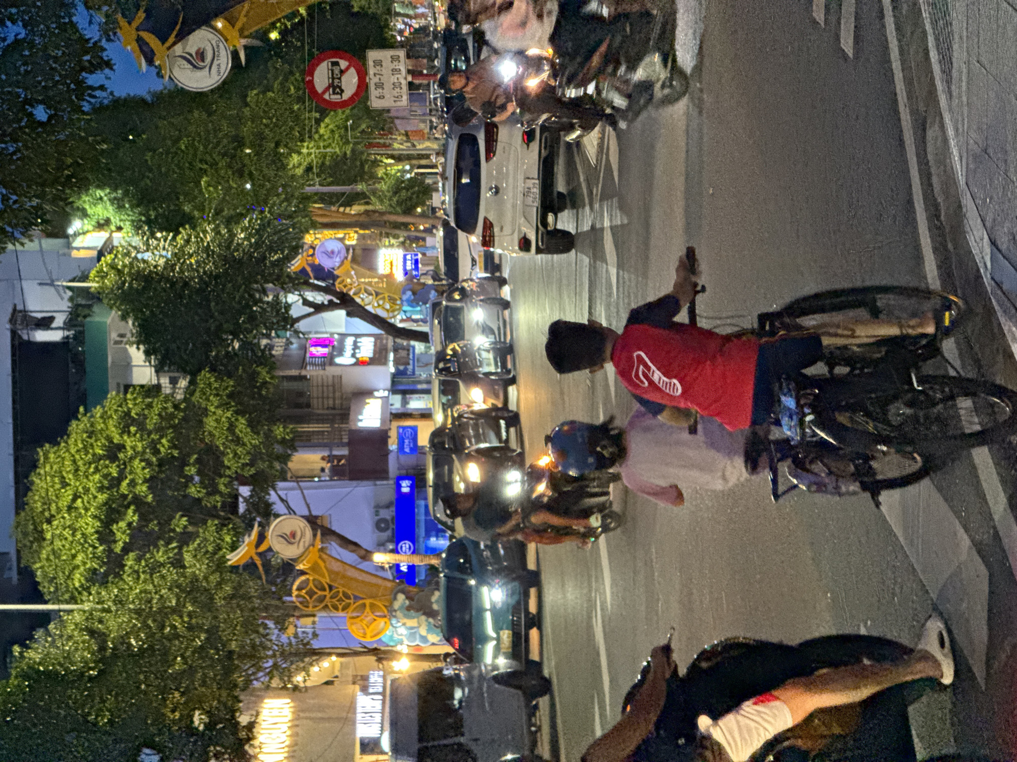 The image depicts a nighttime street scene in Nha Trang, Vietnam, likely a busy intersection or thoroughfare. The street is filled with various vehicles, including cars and motorcycles, illuminated by streetlights and vehicle headlights. In the foreground, two children are on bicycles, one in a red shirt with the number 7 on the back, and the other in a light-colored shirt. They appear to be moving with the flow of traffic. Several motorcycles are also visible, some with riders. The background shows buildings with illuminated signs and trees lining the street, suggesting an urban environment. A traffic sign indicating prohibited hours for vehicles (6:30-7:30 and 16:30-18:30) is visible, further confirming a regulated urban setting. The overall impression is one of a vibrant, active city at night.