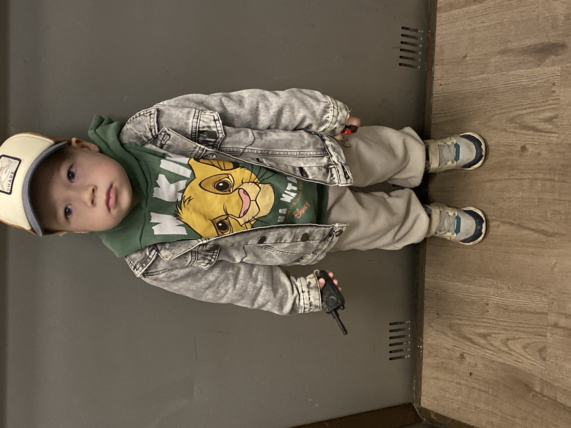 A young child is standing indoors next to what appears to be a utility box or cabinet. The child is wearing a light-colored baseball cap, a green hooded sweatshirt with a "Lion King" graphic, a gray denim jacket, light gray sweatpants, and sneakers. The child is holding a black walkie-talkie in their right hand. The floor is a light wood-grain laminate. The location is not identifiable as being in Sochi, Russia, based on the image alone.