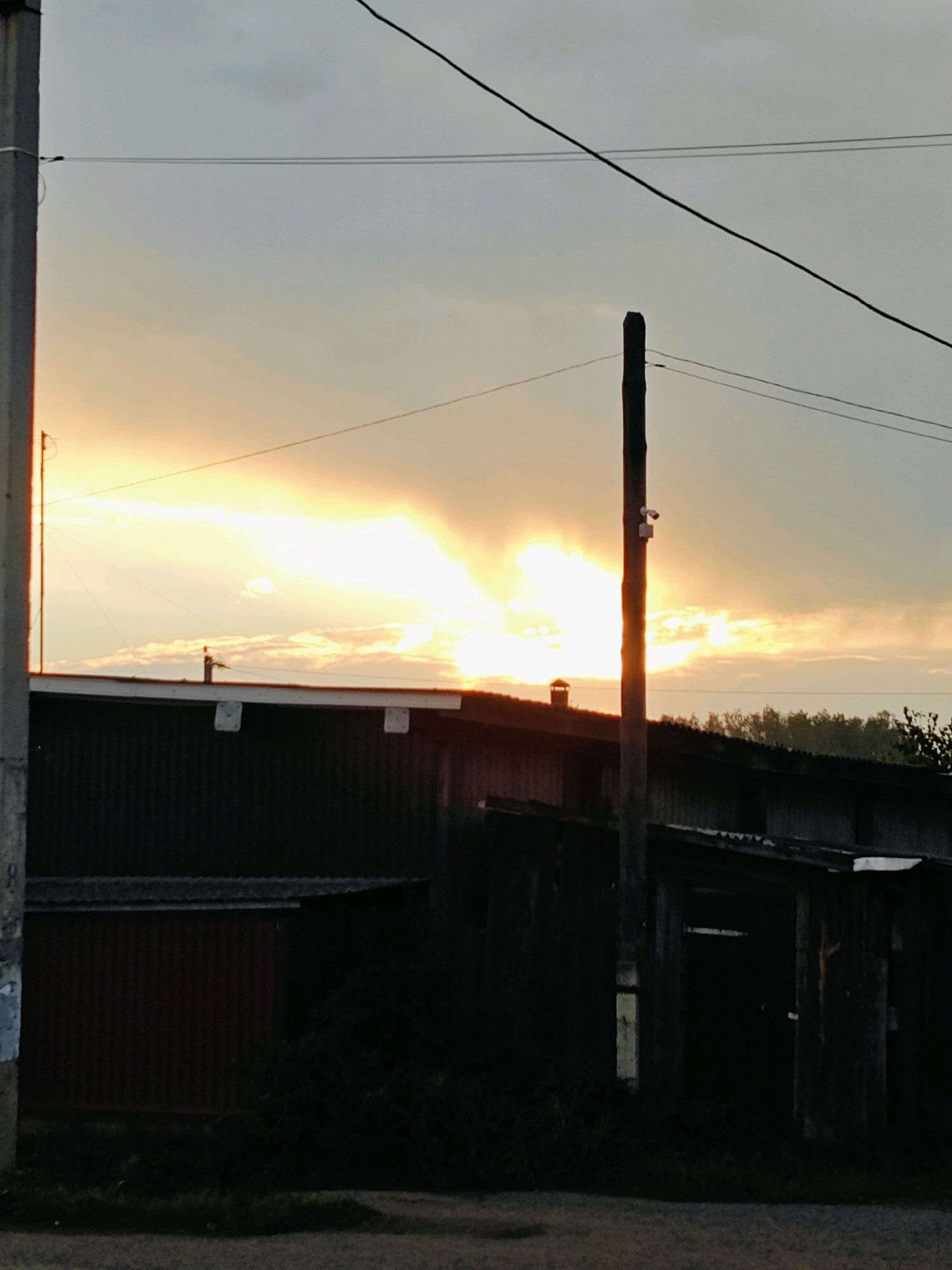 The image depicts a rural scene in the city of Fokino, Russia, at dusk. The sky is filled with dramatic clouds illuminated by the setting sun, which casts a bright, yellowish-orange glow. In the foreground, a dark, corrugated metal building or shed is visible, with a wooden structure attached to its side. A vertical wooden pole, possibly a utility pole, stands prominently in front of the building, with a small security camera mounted on it. Electrical wires stretch across the sky, further indicating the presence of infrastructure. The ground in front of the structures appears to be a dirt or gravel surface, with some sparse vegetation growing. The overall atmosphere is one of a quiet, possibly industrial or agricultural, setting at the end of the day.