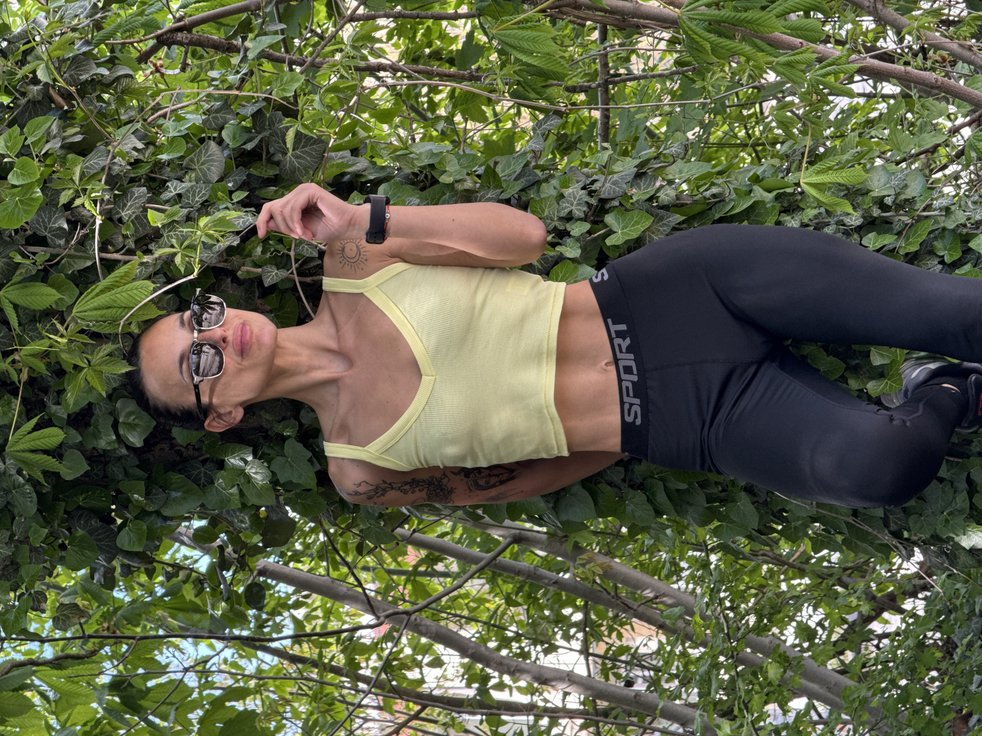 An eye-level shot captures a woman lying amidst dense green foliage in Pleven, Bulgaria. She is looking upwards towards the camera, wearing dark sunglasses that reflect the surroundings, and has her mouth slightly open, suggesting a relaxed expression. Her dark hair is pulled back, and she is dressed in athletic wear: a light yellow ribbed cropped top that exposes her midriff, and black leggings.
A dark-colored smartwatch is visible on her left wrist. She has several tattoos: a stylized sun and crescent moon design on her left shoulder/upper arm, and an intricate pattern on her right forearm. The white text "SPORT" is clearly visible on the black waistband of her leggings. One of her hands is raised, gently holding a small leaf or twig from the surrounding greenery.
The background is dominated by a variety of lush green leaves and tree branches, including distinct ivy foliage, creating a vibrant, natural setting. The bright, natural lighting indicates it is daytime, likely under sunny conditions. The scene suggests a casual outdoor moment or a posed photograph within a park or garden environment.