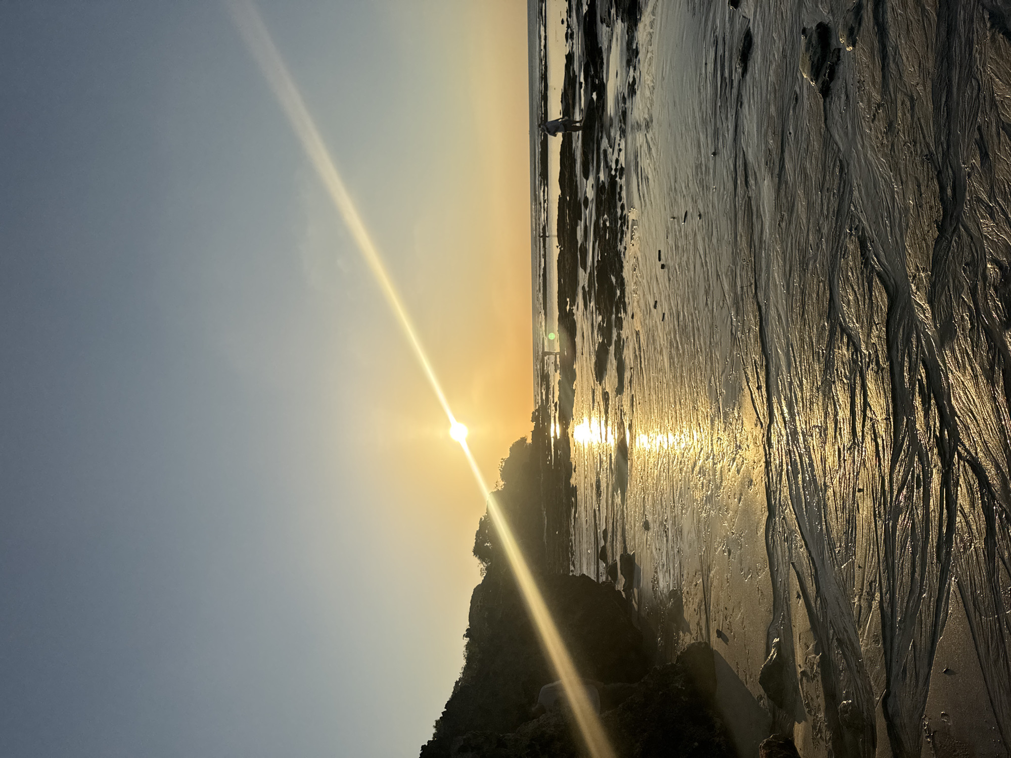 This is a vertical, eye-level, full shot of a beach scene in Kuta Selatan, Indonesia during sunset. The sun is low on the horizon, casting a warm glow and a prominent sunbeam across the sky and water. The foreground consists of wet sand with visible ripple patterns, indicating the receding tide. Rocky outcrops are scattered across the beach and extend into the shallow water. In the distance, the ocean meets the sky, with a few small figures visible on the water's edge and in the shallow areas. The sky is a gradient of soft blue and orange hues. There are no signs of any infrastructure or buildings visible in the frame.