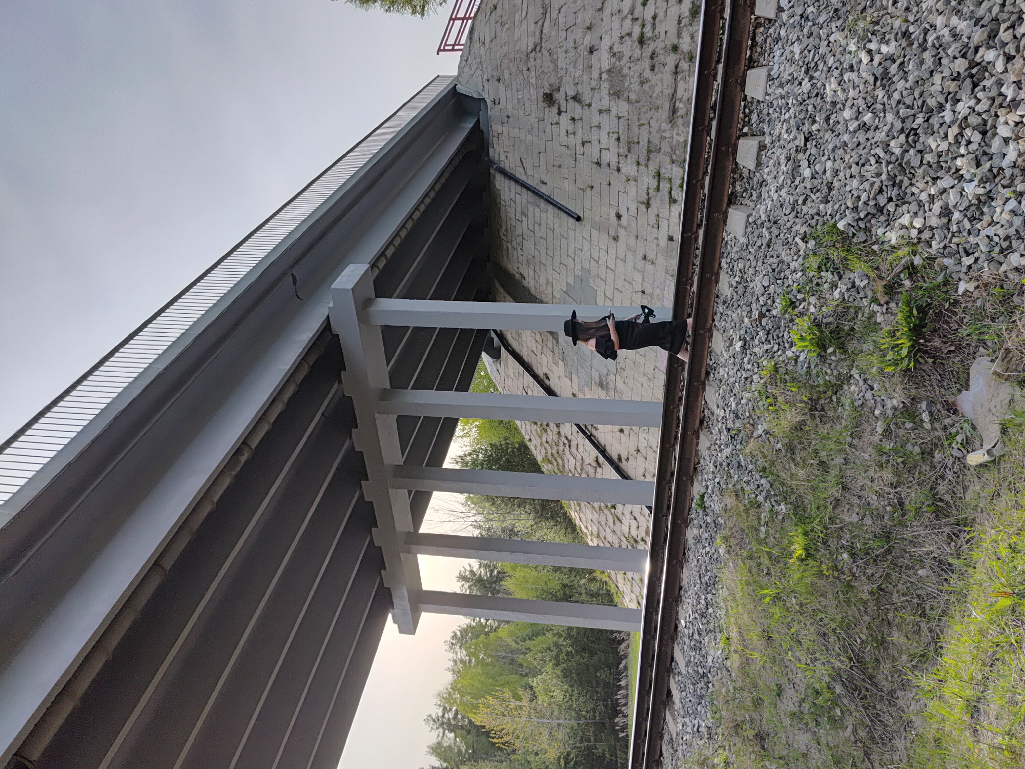 A woman in a black outfit and hat stands on a set of train tracks. She is next to a concrete platform and underneath a concrete overpass. The train tracks are surrounded by gravel and vegetation. The location is Chaikovsky, Russia.