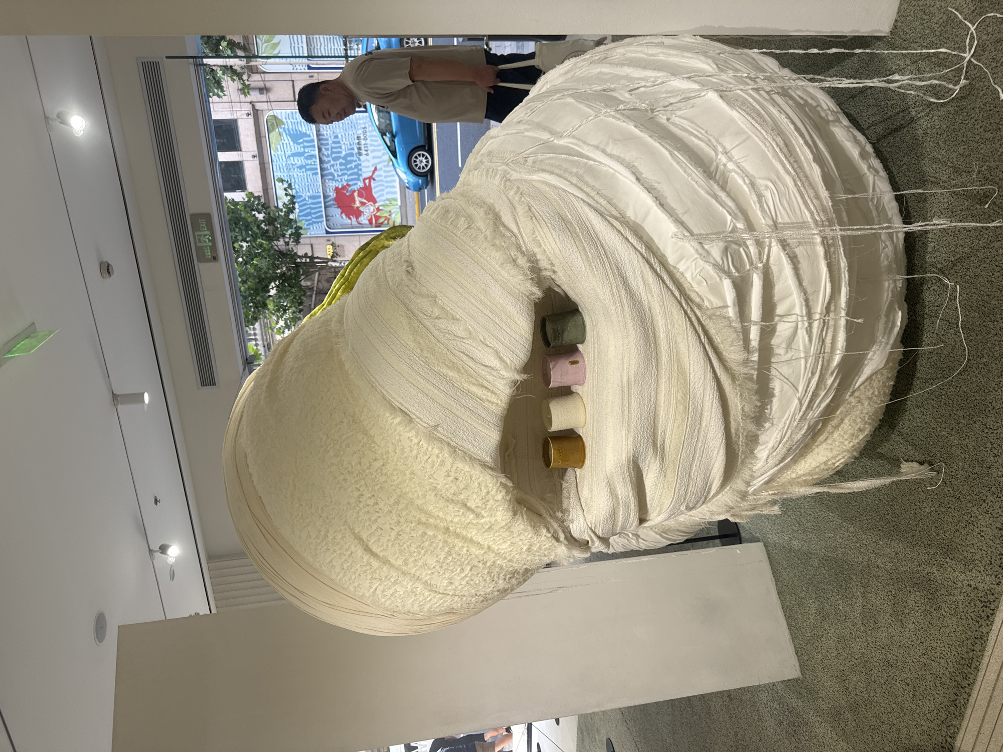 A man stands near a large, abstract sculpture made of white fabric in what appears to be a store or gallery. The sculpture is spherical with layered, textured fabric, and has an opening in the center where several small cylindrical objects are placed. The man is looking to his right, away from the sculpture. Behind him, through a large window or doorway, a street scene is visible with a blue car, trees, and buildings. An "EXIT" sign is also visible above the opening. The floor is carpeted with a speckled grey pattern. The lighting suggests an indoor environment with artificial lights.