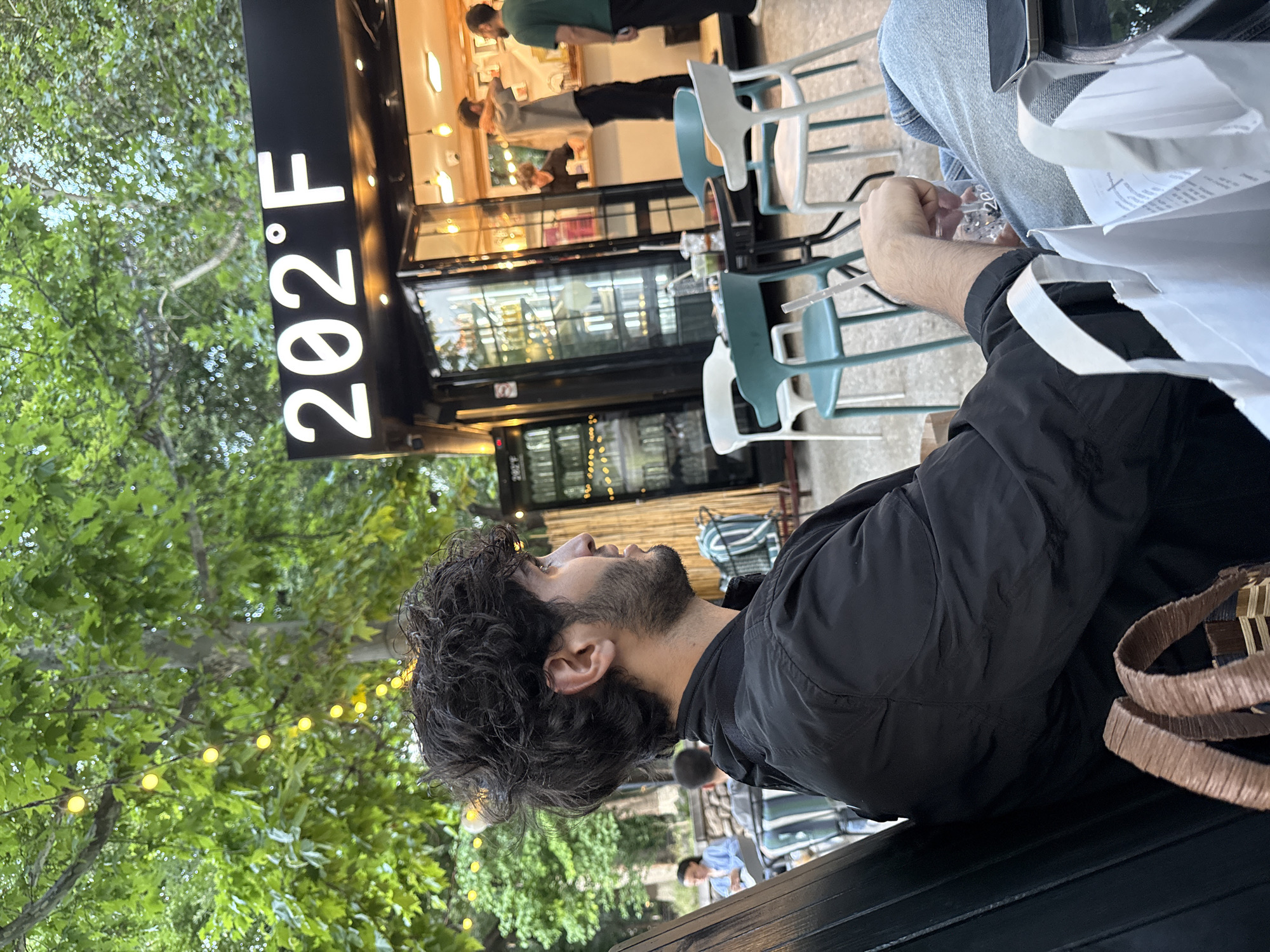 The image depicts a man sitting outdoors at a cafe. He is wearing a black jacket and jeans, and has dark, curly hair and a beard. He is holding a drink with a straw and looking to his right. In the background, there is a food truck or kiosk with the sign "202°F" and string lights overhead. Several people are visible inside and around the kiosk, and there are outdoor tables and chairs. The environment appears to be a public space with trees and foliage. The location is identified as Yerevan, Armenia.