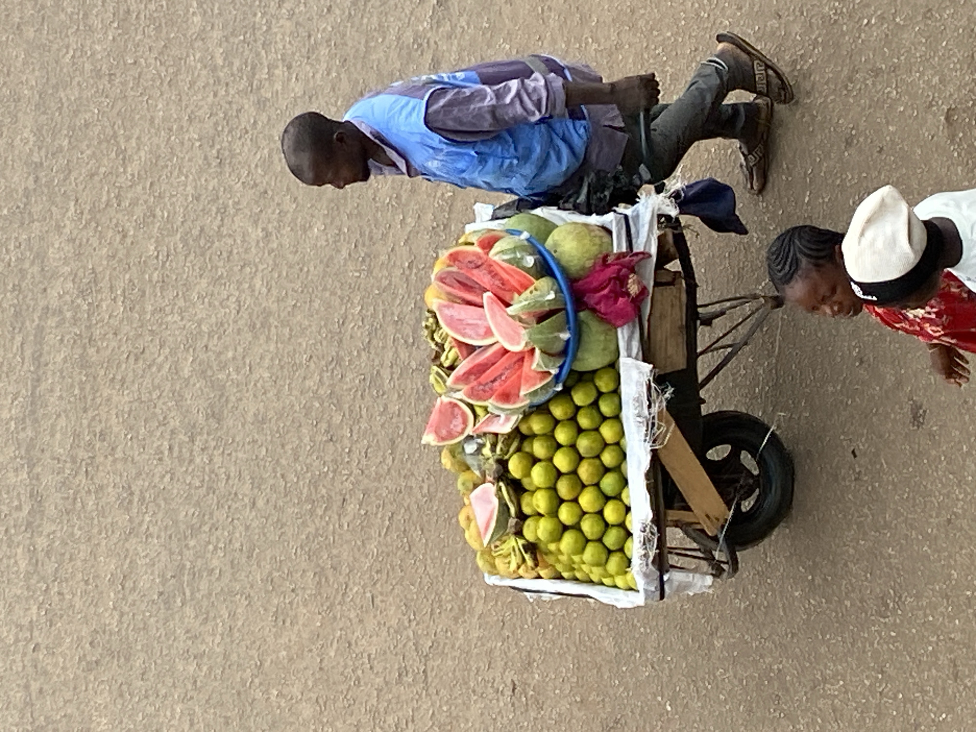 An aerial view captures a scene in Bukuru, Nigeria. A man pulls a cart laden with produce. The cart contains watermelons and what appear to be limes. A woman, standing adjacent to the cart, seems to be engaged with the vendor. They are on a sandy, open area.