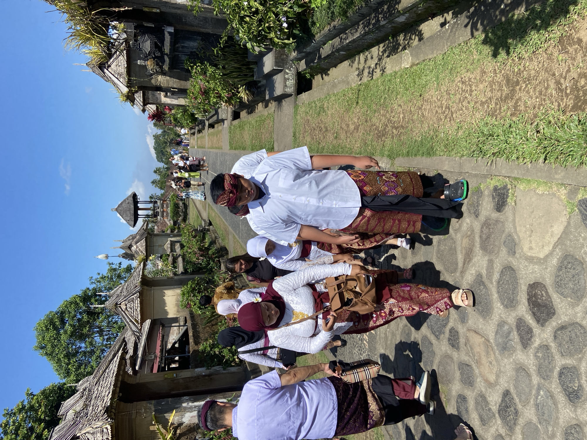 The image depicts a group of people walking up a stone pathway in what appears to be a temple complex. The architecture suggests a Southeast Asian style, possibly Balinese, with ornate roofs and stone carvings. The people are dressed in traditional clothing, with men wearing sarongs and women in headscarves and long skirts. The pathway is lined with lush greenery and stone structures. In the background, more people can be seen walking further up the path, and a clear blue sky with a few scattered clouds is visible. The scene conveys a sense of cultural pilgrimage or tourism. Based on the architectural style and attire, the location is likely the Kehen Temple in Bangli, Indonesia, known for its impressive banyan tree and ancient stone inscriptions.
