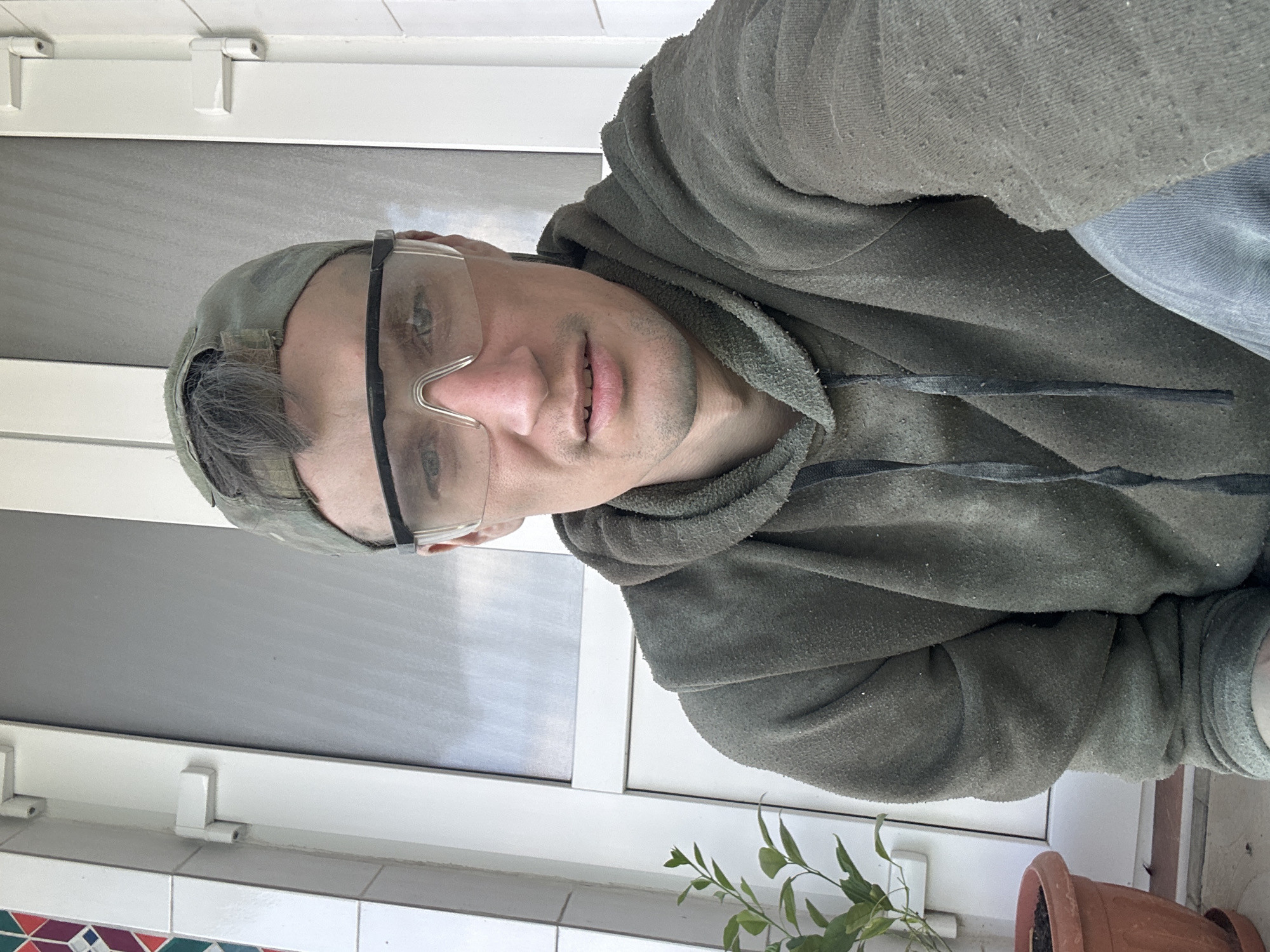 The image is a selfie taken by a man wearing a camouflage baseball cap, clear safety glasses, and a dark green hooded sweatshirt. He is looking directly at the camera with a neutral expression. The background appears to be an exterior setting, possibly a porch or balcony, with a white door and window visible. To the left of the man, there is a small potted plant with green leaves. The man's clothing has some fuzzy texture, and there are small specks on it, suggesting it might be dusty or from a workshop. The lighting suggests it is daytime, with natural light illuminating the scene. The man appears to be in a casual or work-related environment. The provided location of Makiv, Ukraine, does not offer specific contextual details relevant to the visual elements in the image itself.